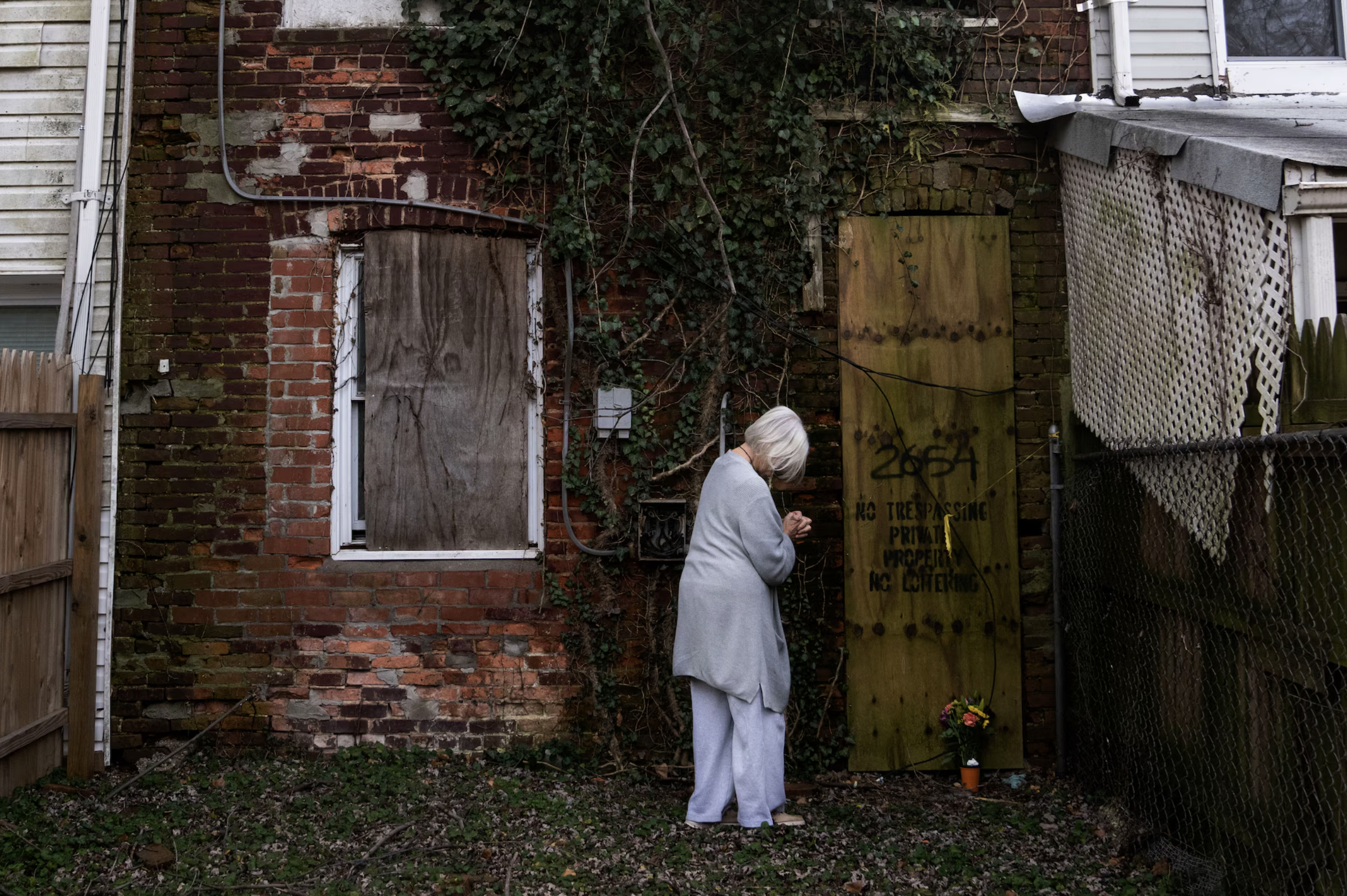 An elderly woman with white hair, wearing a gray sweater, stands in front of a backyard brick wall covered with ivy, looking down while holding something in her hands. There is a large yellow 'No Trespassing' sign on the wall. To her right, there is a flower pot with bright flowers on the ground.
