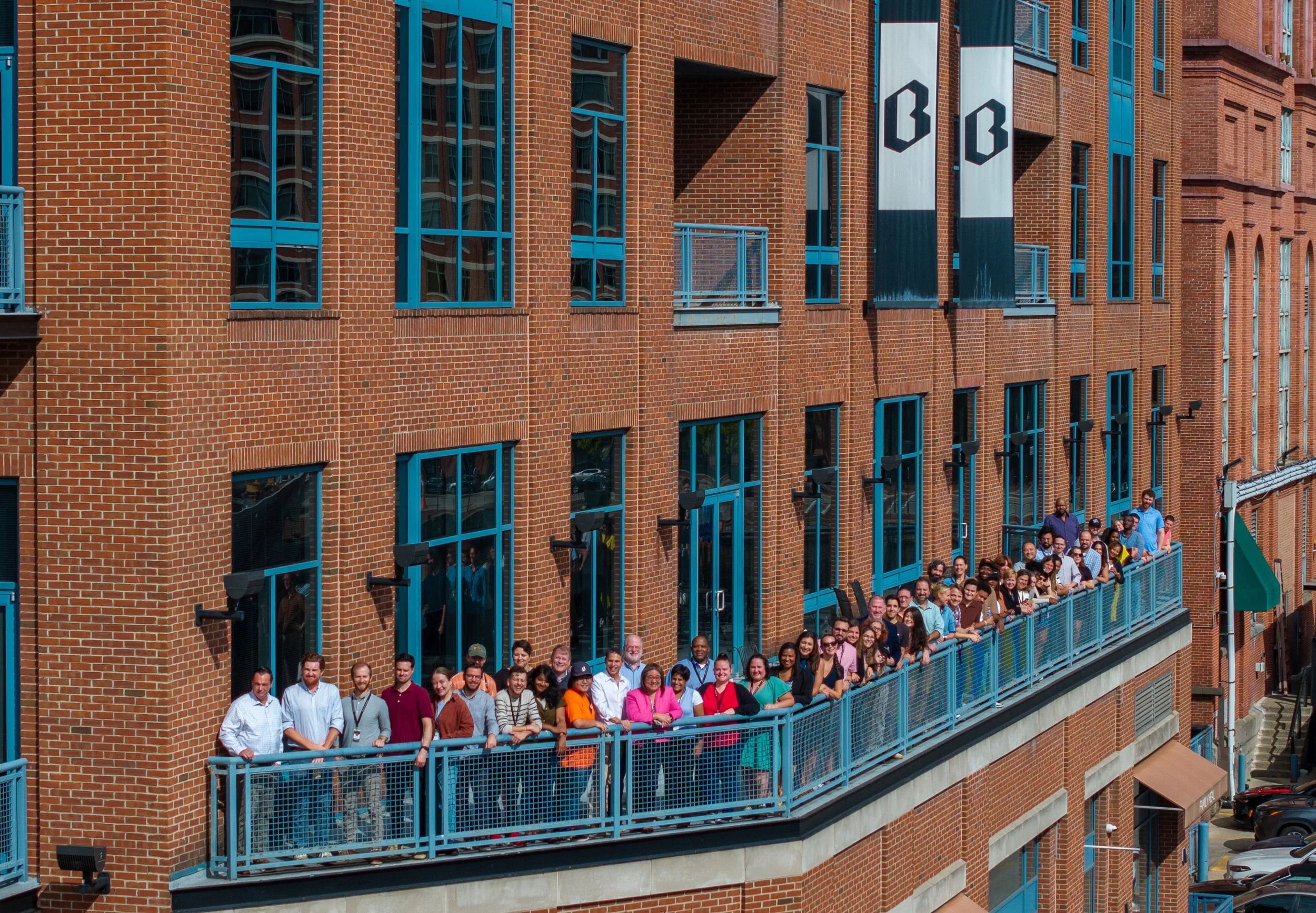 A group of people standing on a blue balcony outside a brick building with large windows and banners displaying the letters 'B B'.