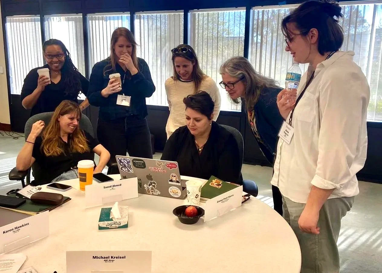 Group of women gathered around a table, looking at a laptop together, some holding coffee cups and water bottles, in a bright room with large windows.