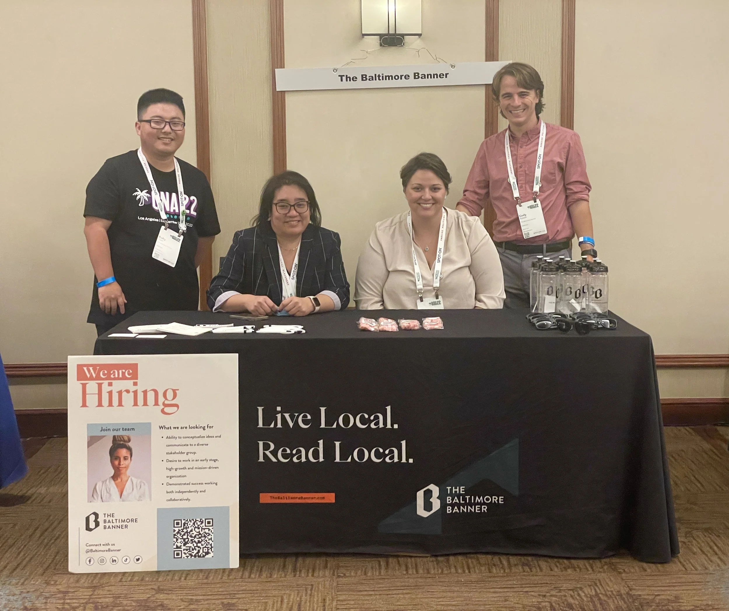 A group of four people standing at a table with promotional materials, with a sign behind them reading 'The Baltimore Banner'. The table displays a large sign reading 'We are Hiring' and encourages reading local news. There are water bottles and pens on the table, and the individuals are wearing lanyards and name tags.