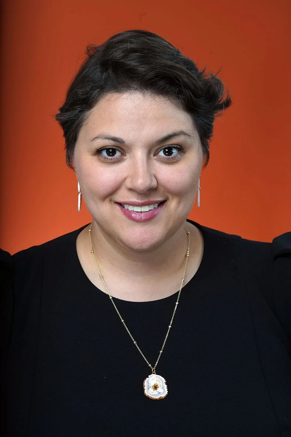 A woman with short dark hair and fair skin smiling, wearing a black top, drop earrings, and a gold necklace with a white and gold pendant, against an orange background.