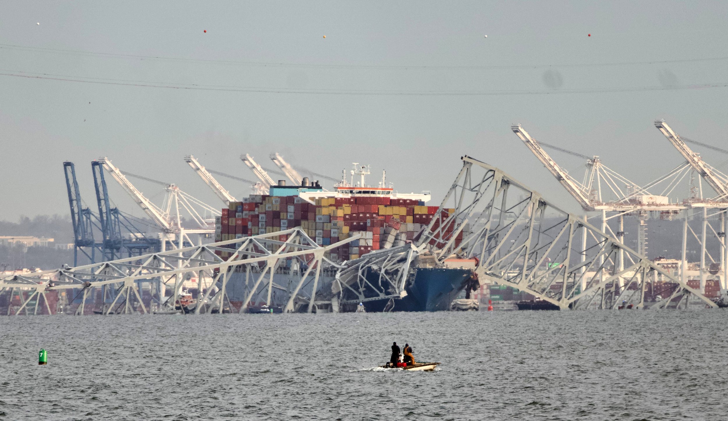 A large container ship with stacked containers is sinking after a collision, with debris and broken structures in the water. A small boat with people is nearby in the foreground.