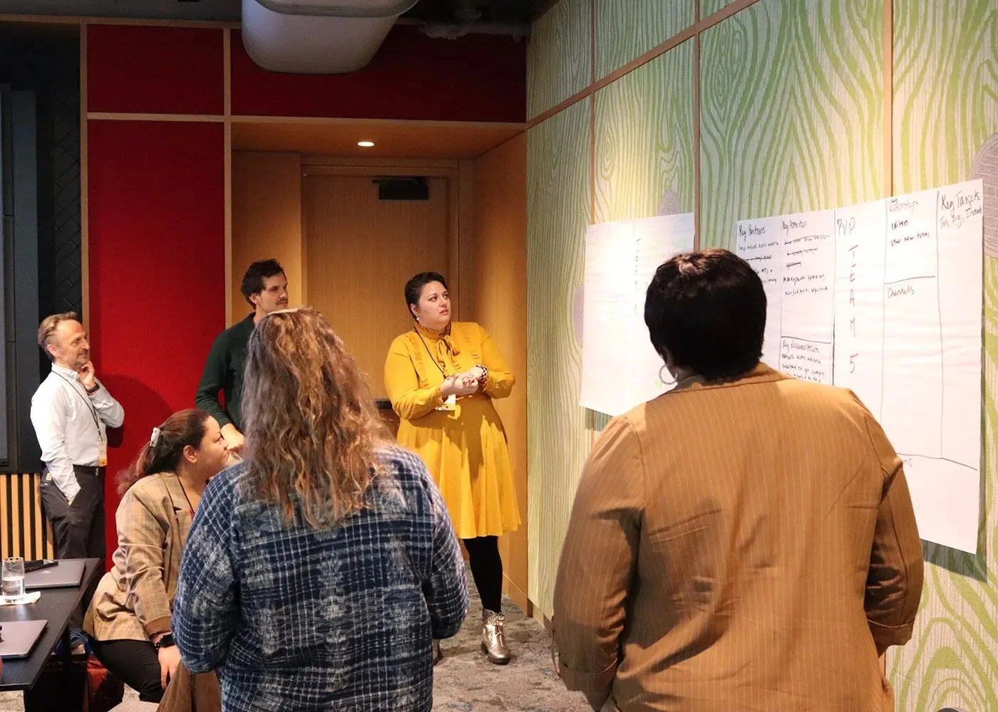 Group of people attending a presentation or workshop, with some taking notes and one woman in a yellow dress speaking in front of a wall with papers or charts.
