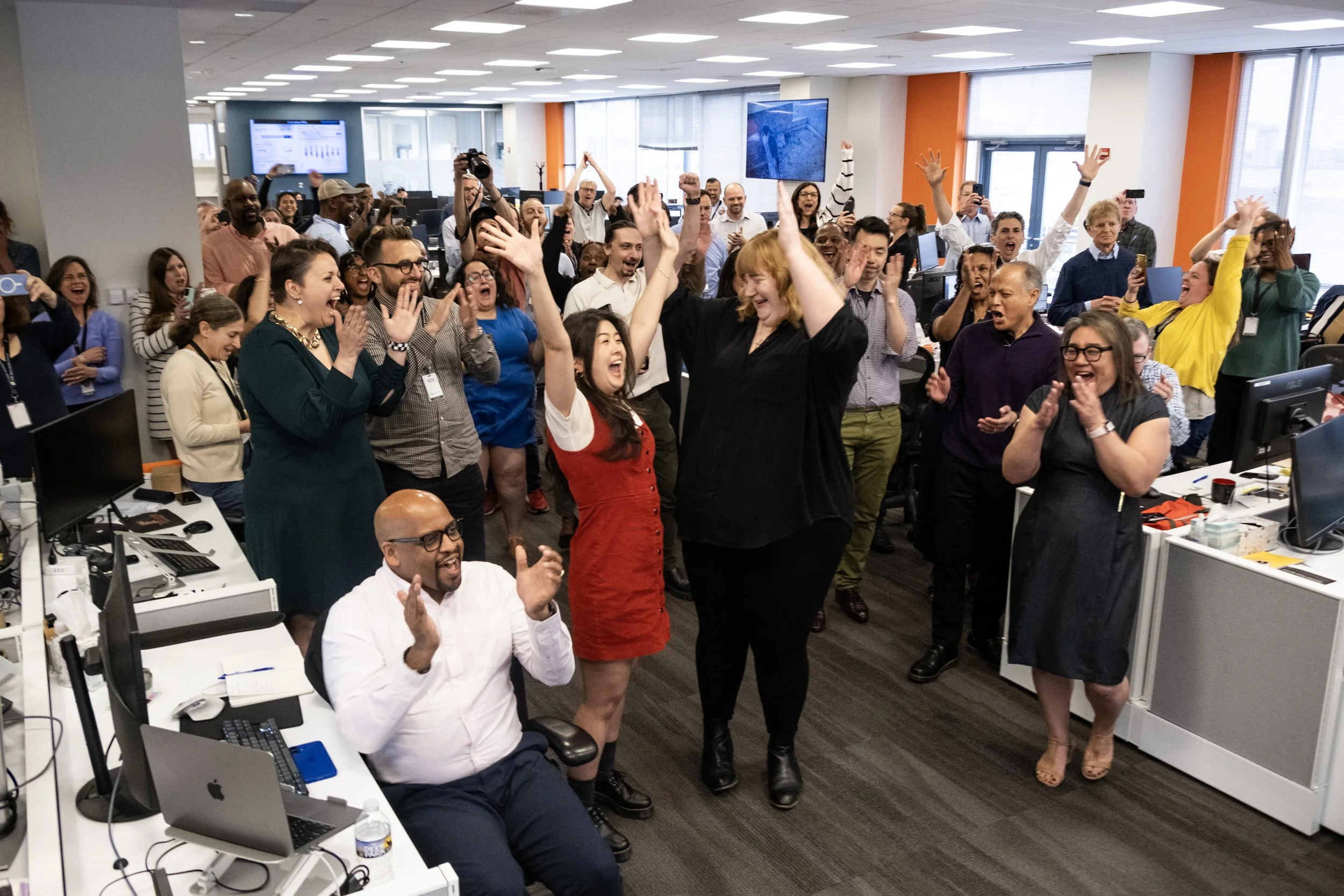 A large group of diverse people in an office celebrating, cheering, and clapping with some raising their hands in the air. People are smiling, and some are taking photos. The office has many desks with computers, large windows, and screens on the wal