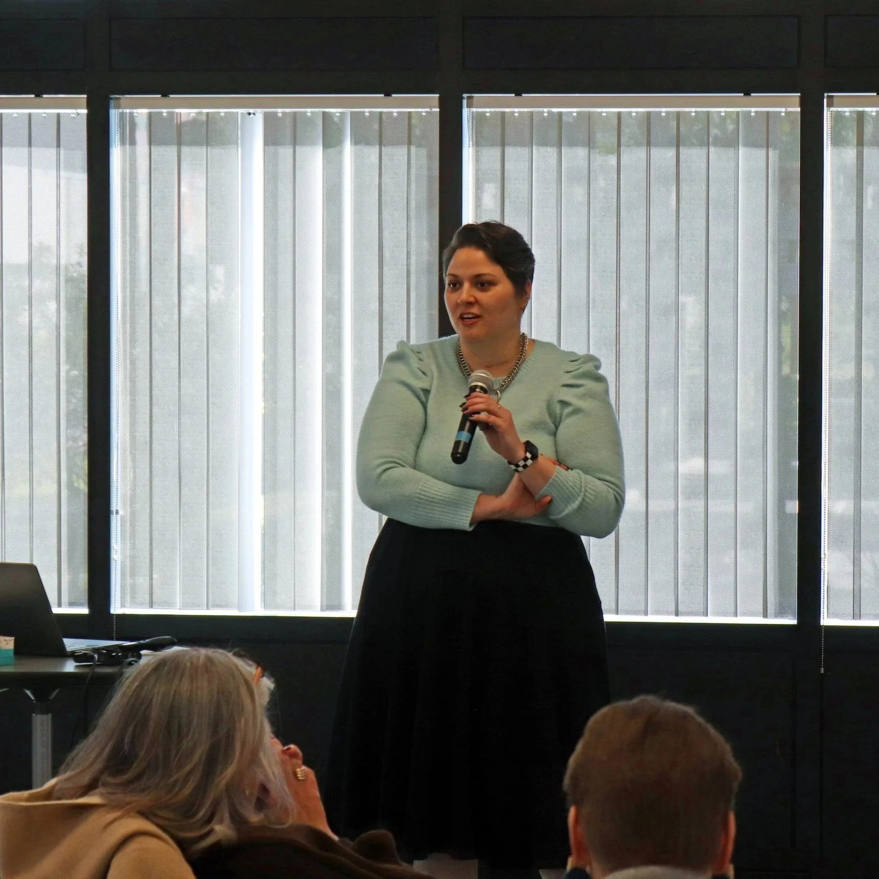 A woman with short dark hair, wearing a light blue sweater and a chunky necklace, is speaking into a microphone in front of large window blinds. Audience members are visible in the foreground.