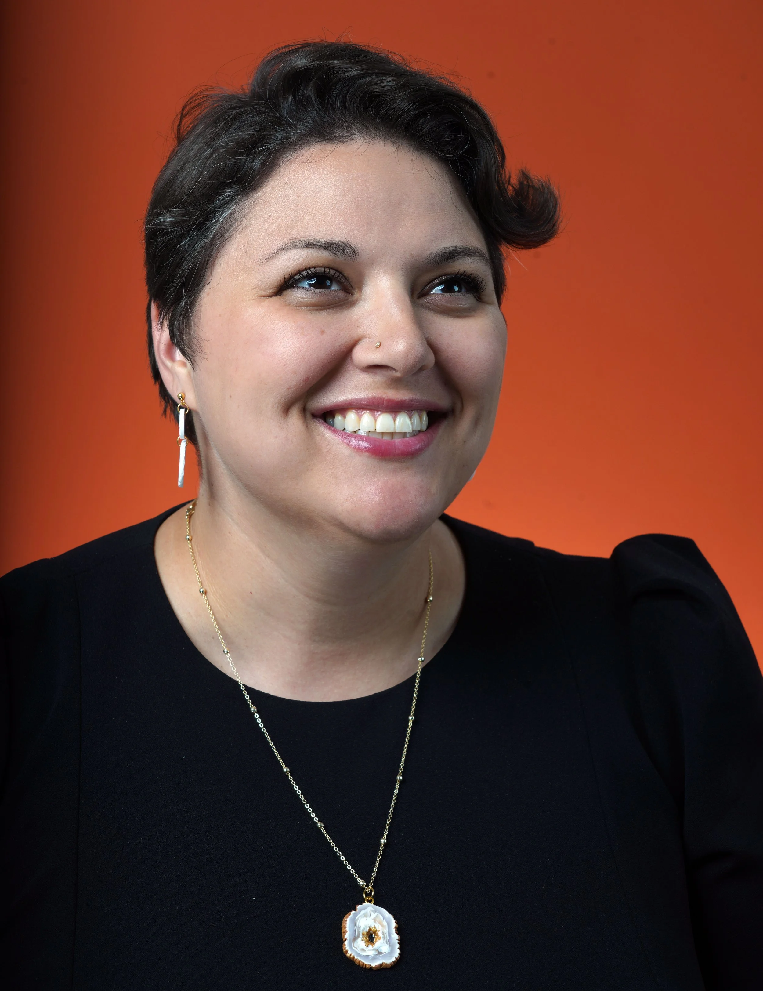 A woman with short dark hair, hoop earrings, wearing a black top, and a gold necklace with a large floral pendant, smiles against an orange background.