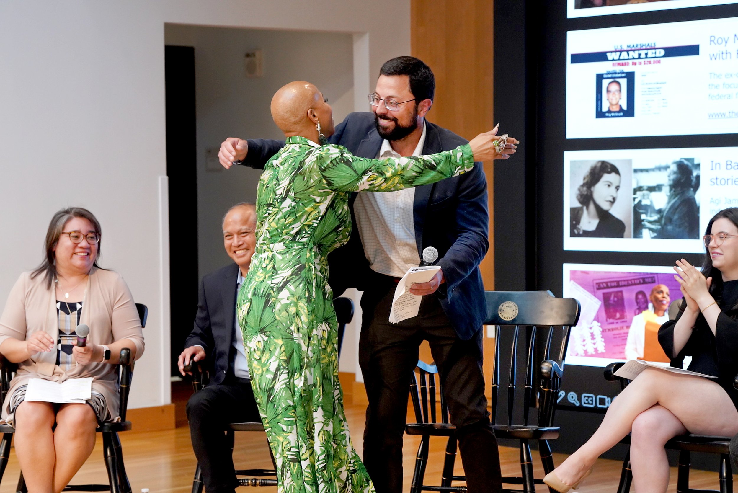 A group of people participating in a panel or discussion, with a woman in a green leafy dress hugging a man in a dark suit. Other seated women and men are visible, some smiling or holding microphones, and a large screen with images and text is in the background.