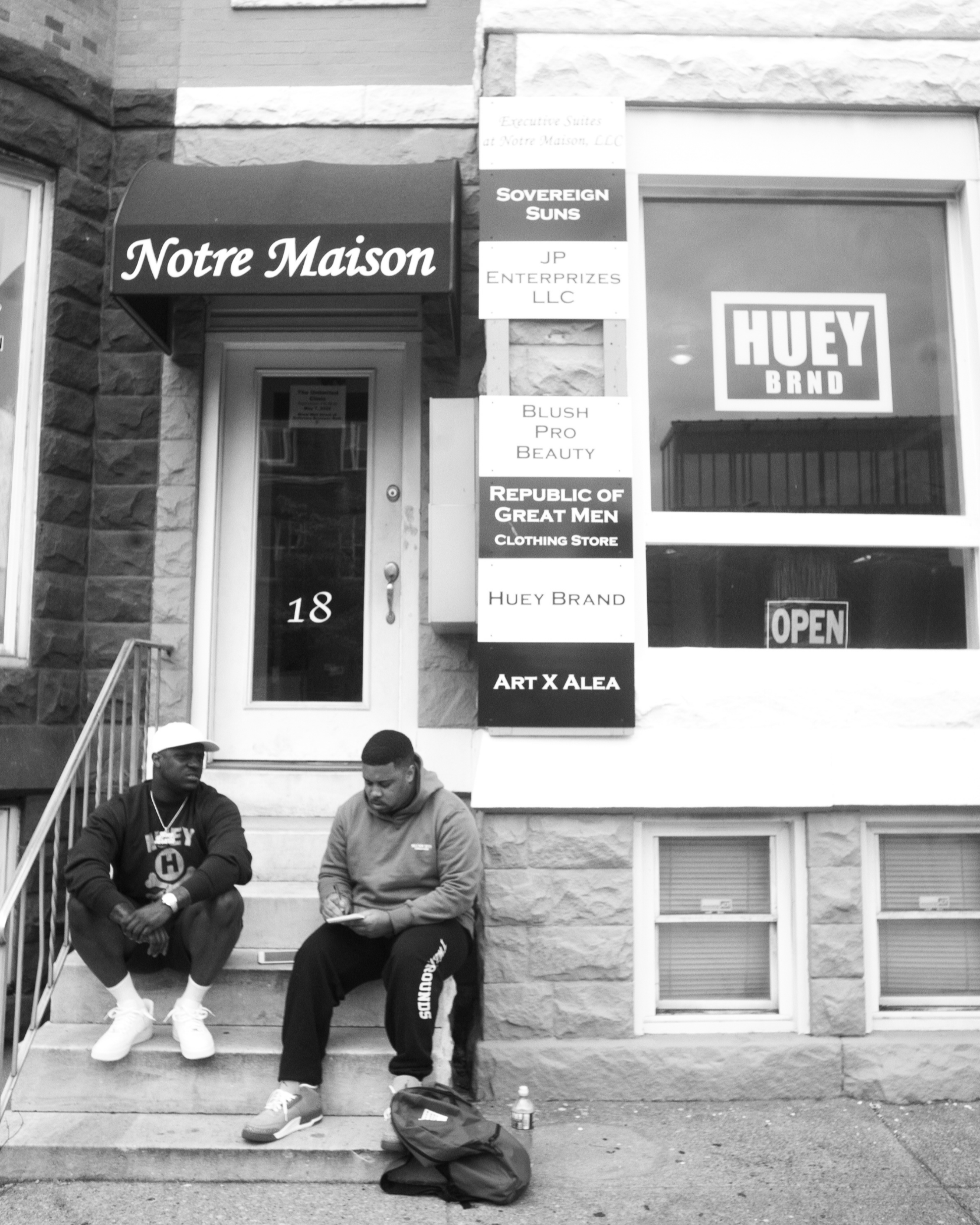 Two young men sitting on steps outside a building, one wearing a white cap and hoodie, and the other reading a paper. A backpack and water bottle are on the ground nearby. The building has a brick facade, windows with blinds, and several business signs including 'Notre Maison,' 'Sovereign Suns,' 'Huey Brand,' and 'Art X Alae'.