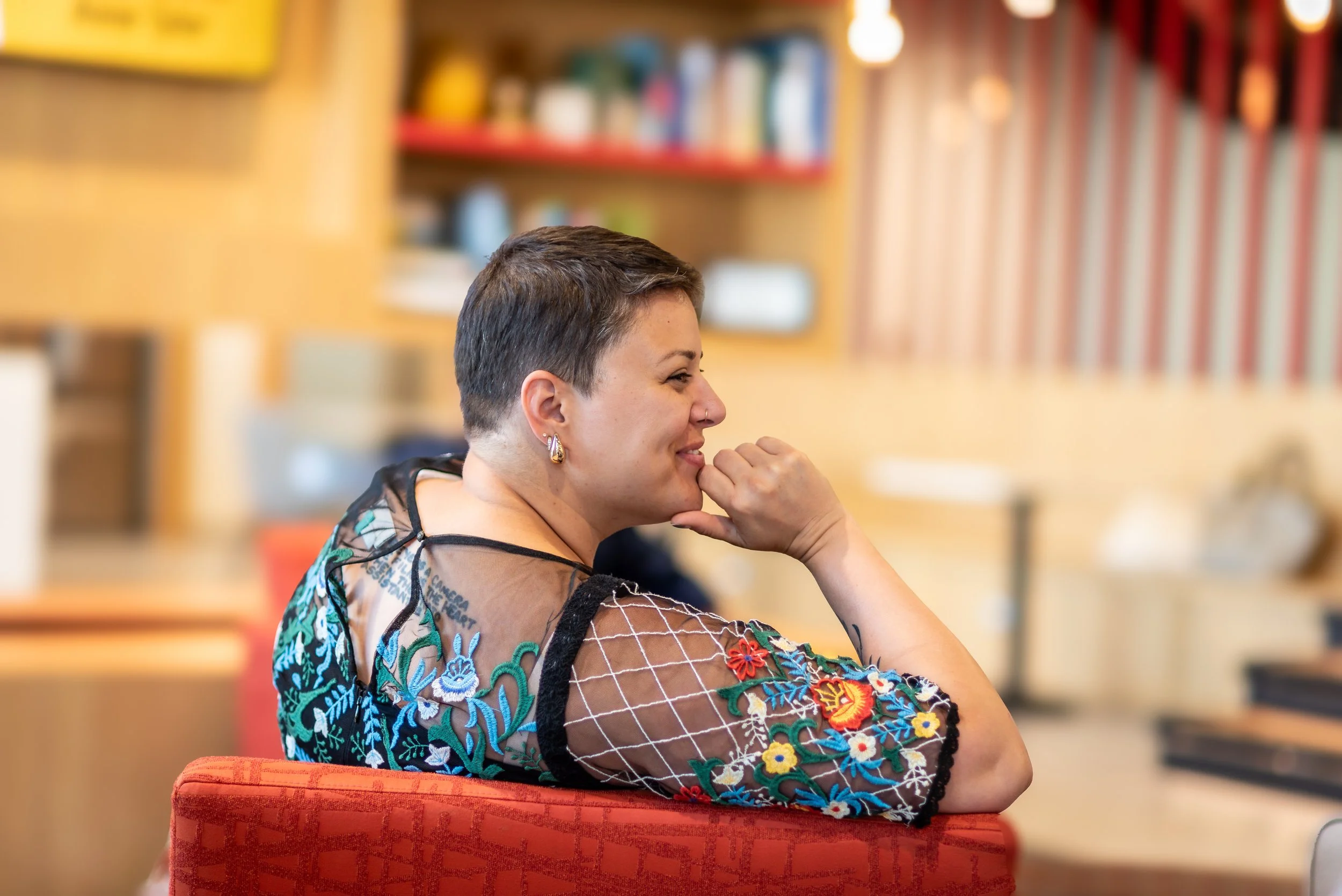 A woman with short hair in profile sitting on an orange chair, smiling with her hand near her mouth in a colorful room with bookshelves.