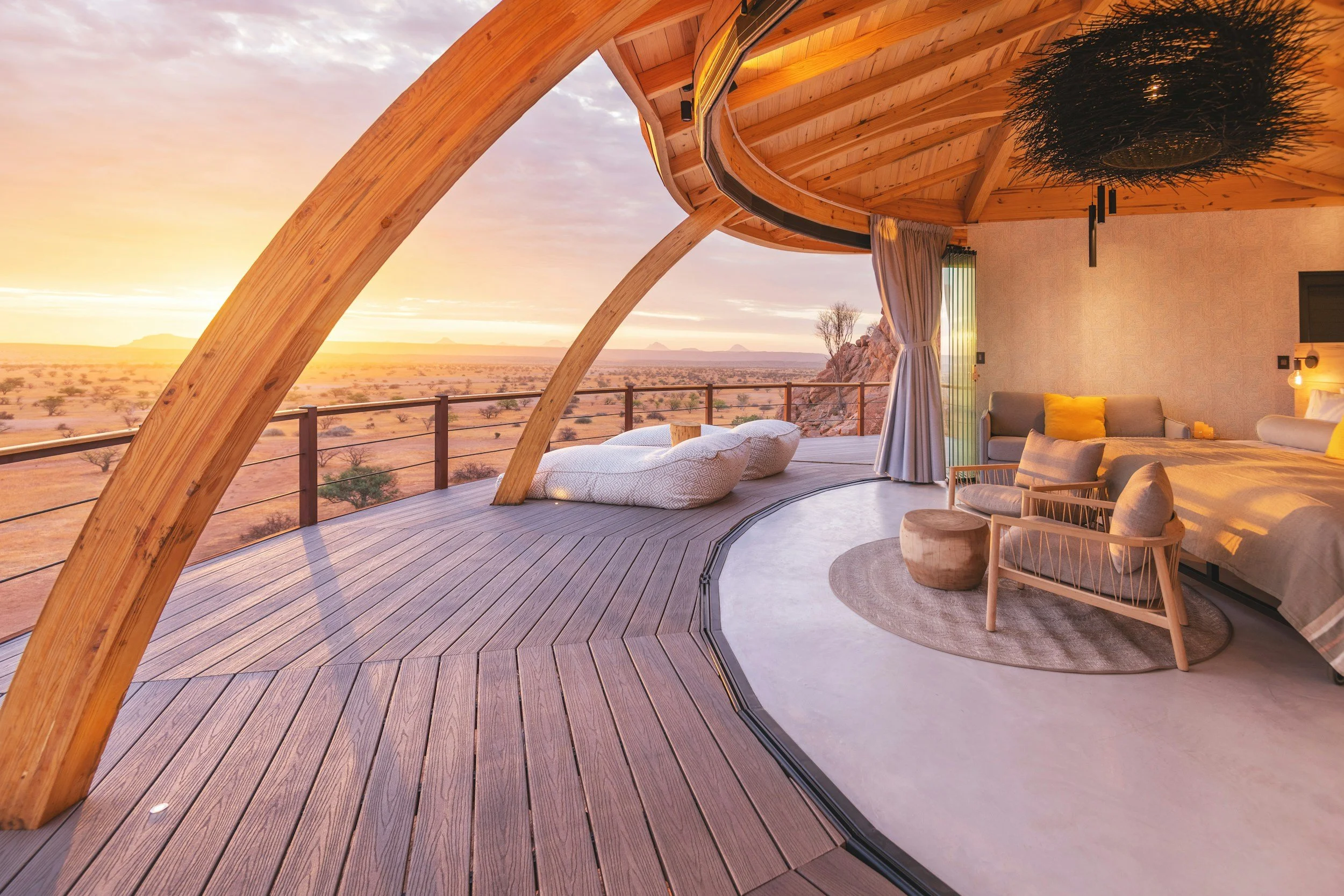 Interior of a modern, curved wooden room with large glass windows overlooking a desert landscape at sunset. The room has floor seating with cushions, a small wooden table, chairs, and a hanging plant.
