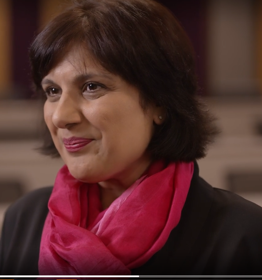A middle-aged woman with short dark hair wearing a black blazer and pink scarf, smiling softly indoors.
