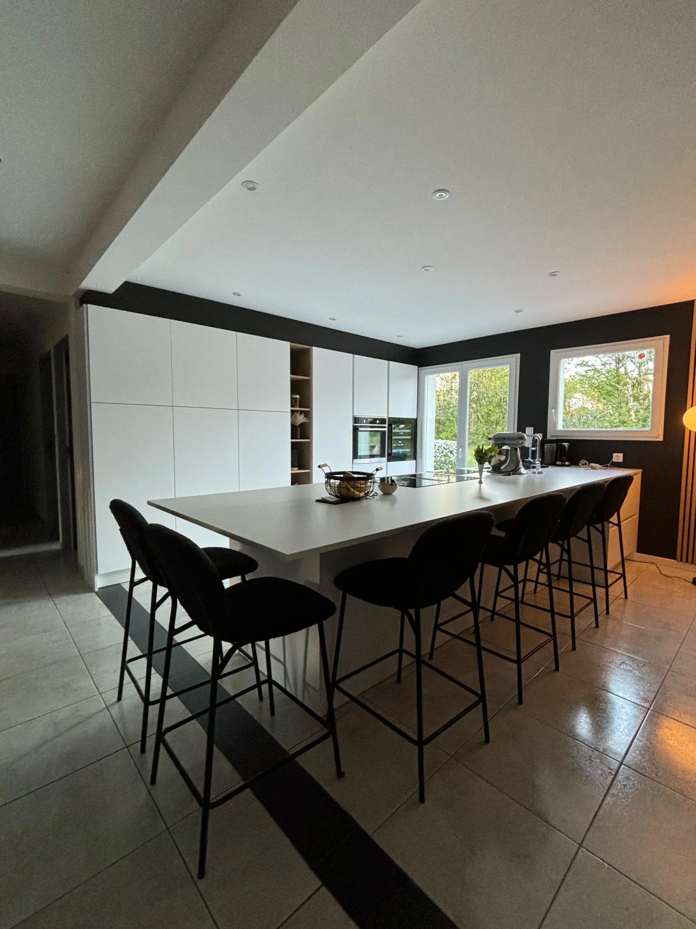 Cuisine moderne rénovée dans la maison à charnay avec îlot central blanc, crédence noire, deux fenêtres laissant entrer la lumière naturelle, et des chaises autour de l'îlot. Présence d'appareils électroménagers modernes.