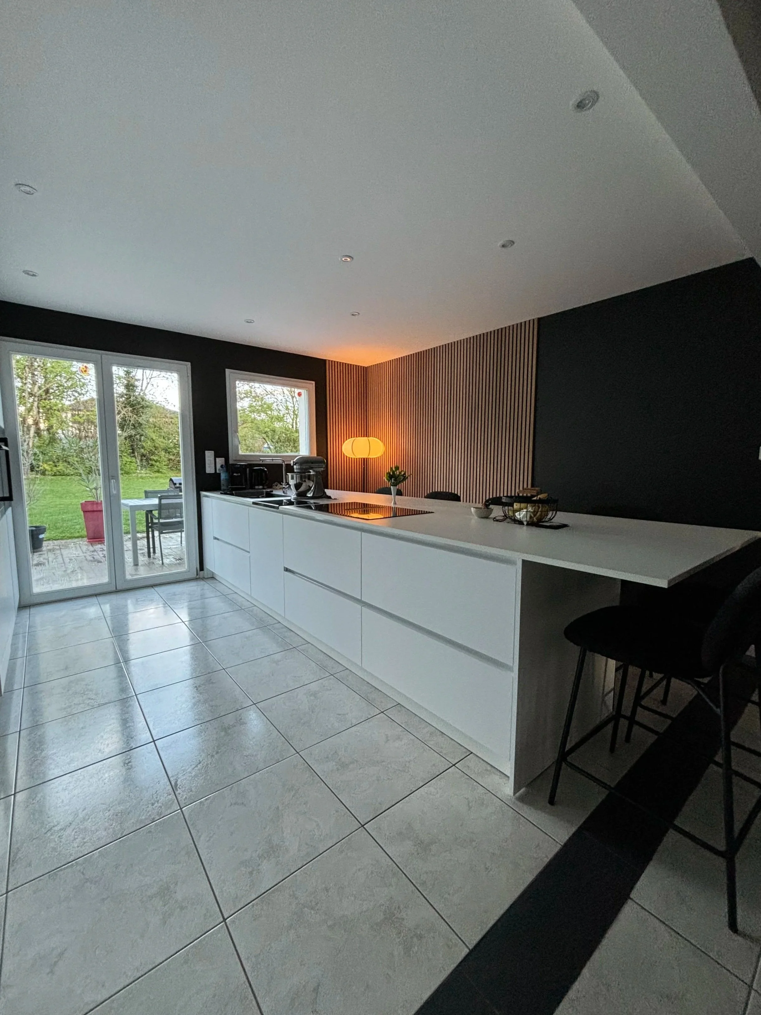 Cuisine moderne rénovée dans la maison à charnay avec plans de travail blancs, fenêtres donnant sur un jardin, lampadaire orange, murs noirs avec une paroi en bois, carrelage gris, quelques objets de cuisine et huit chaises noires.