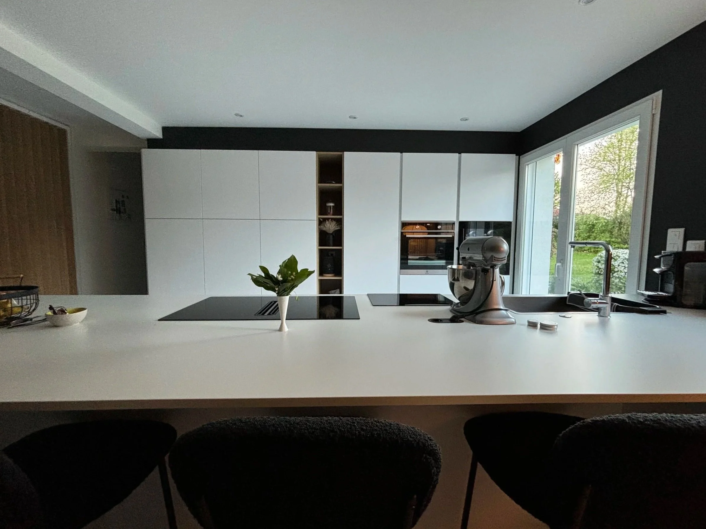 Cuisine moderne rénovée dans la maison à charnay avec des armoires blanches, un plan de travail blanc, une fenêtre donnant sur un jardin, un robot de cuisine sur le plan de travail, et des sièges noirs en premier plan.