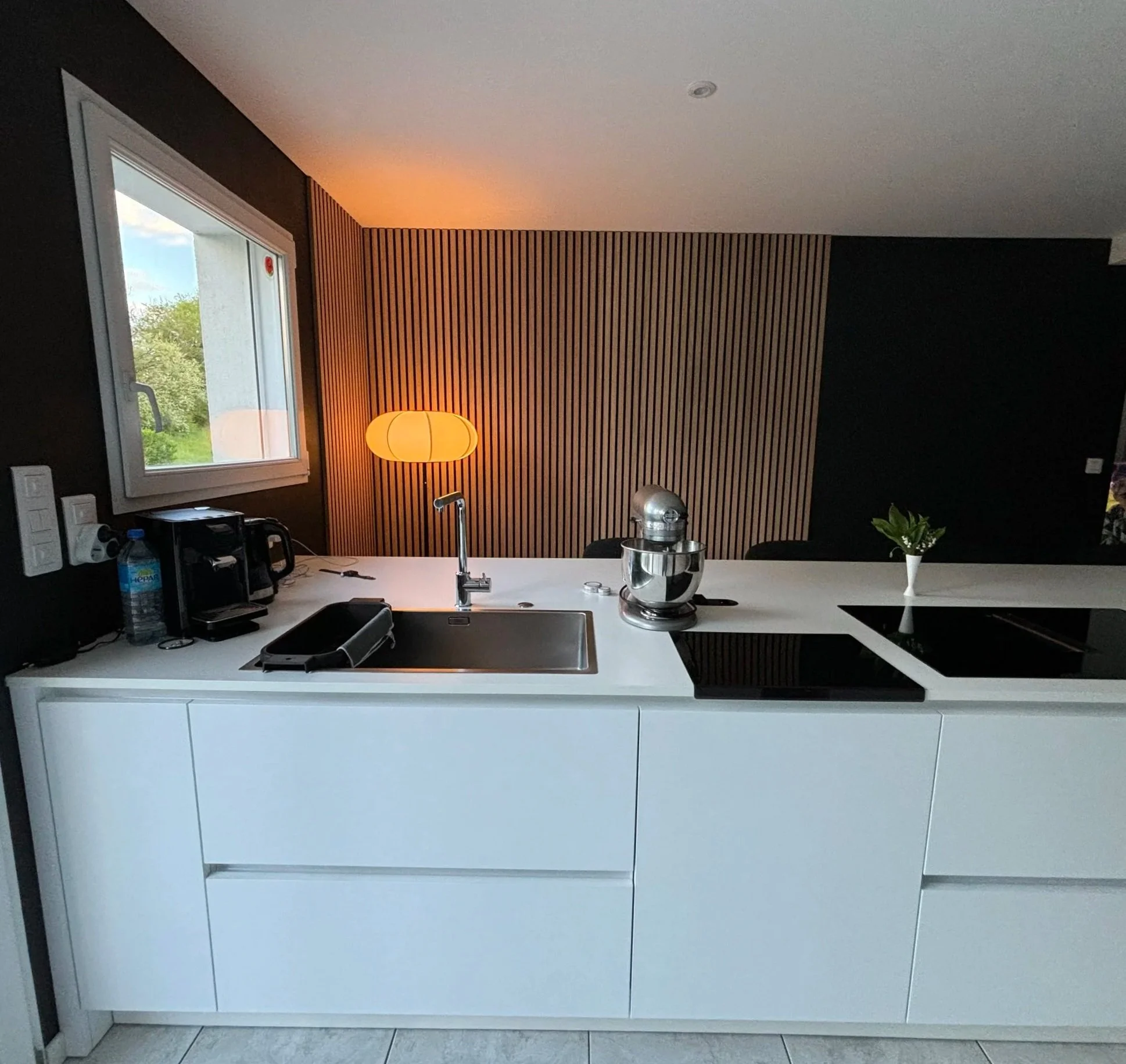Cuisine moderne rénovée dans la maison à charnay avec un plan de travail blanc, un évier, un mixeur, une plante dans un vase blanc, une lampe orange, une fenêtre laissant entrer la lumière naturelle, un mur avec un revêtement en bois.