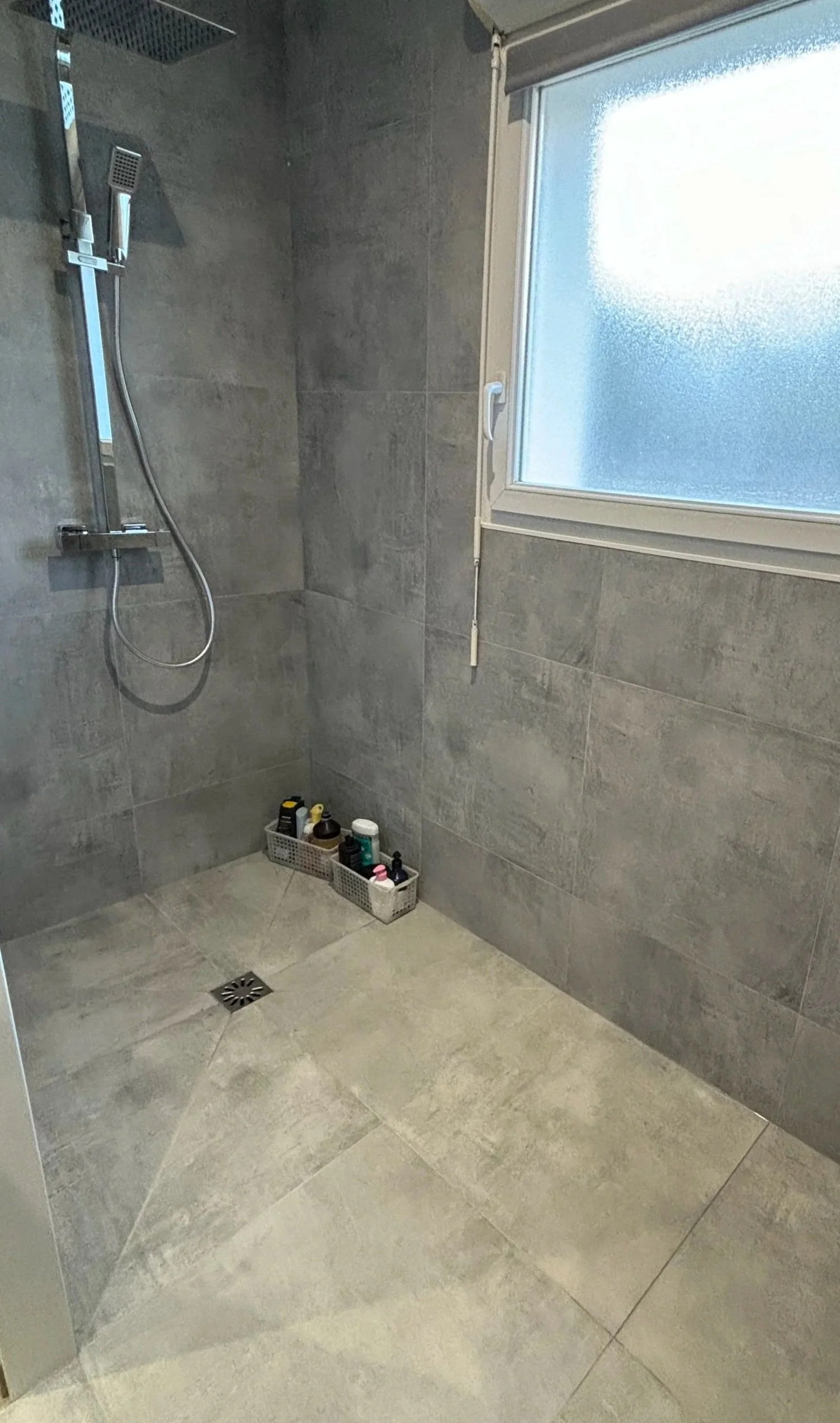 Salle de douche moderne avec grande fenêtre, paroi en carrelage gris, douches à main et étagère contenant divers produits de toilette, sol en carreaux gris, et un drain de sol. Agencement sur mesure. Maison charnay.