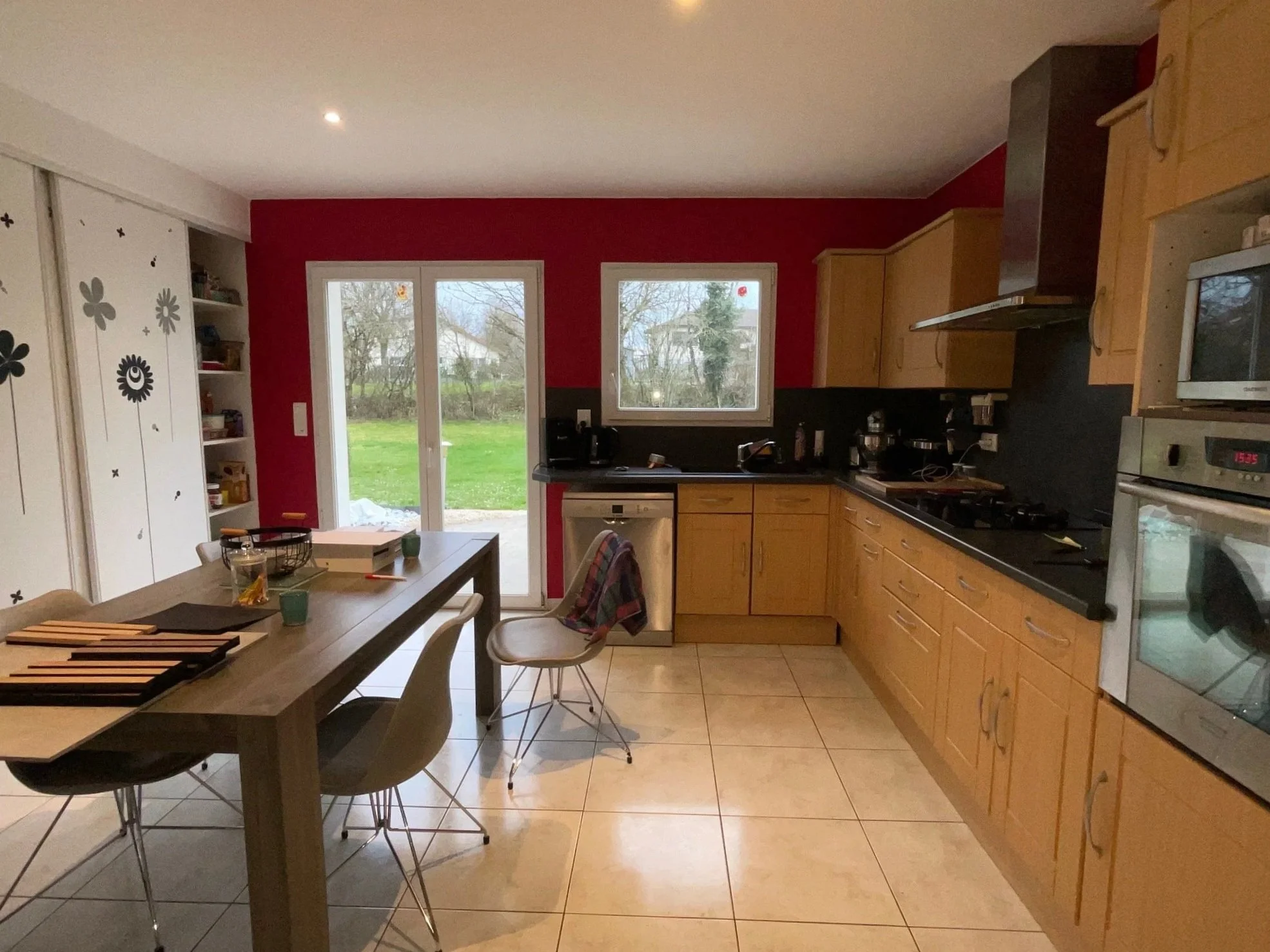 Cuisine existante de la maison rénovée à charnay avec murs rouges, armoires en bois, un plan de travail noir, deux fenêtres donnant sur un jardin et une porte-fenêtre.