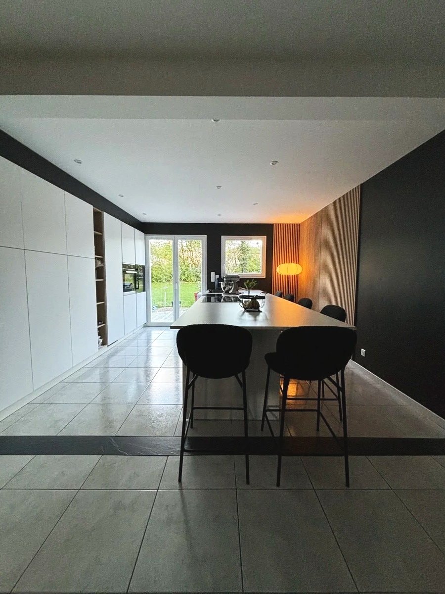 Cuisine rénovée dans la maison à charnay moderne avec un îlot central, chaises, grande fenêtre laissant entrer de la lumière naturelle, mur en bois, lampe sur le mur à droite.