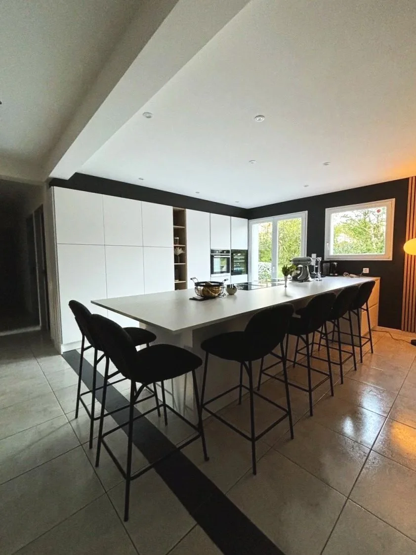 Cuisine moderne rénovée dans la maison à charnay avec îlot central blanc, crédence noire, deux fenêtres laissant entrer la lumière naturelle, et des chaises autour de l'îlot. Présence d'appareils électroménagers modernes.