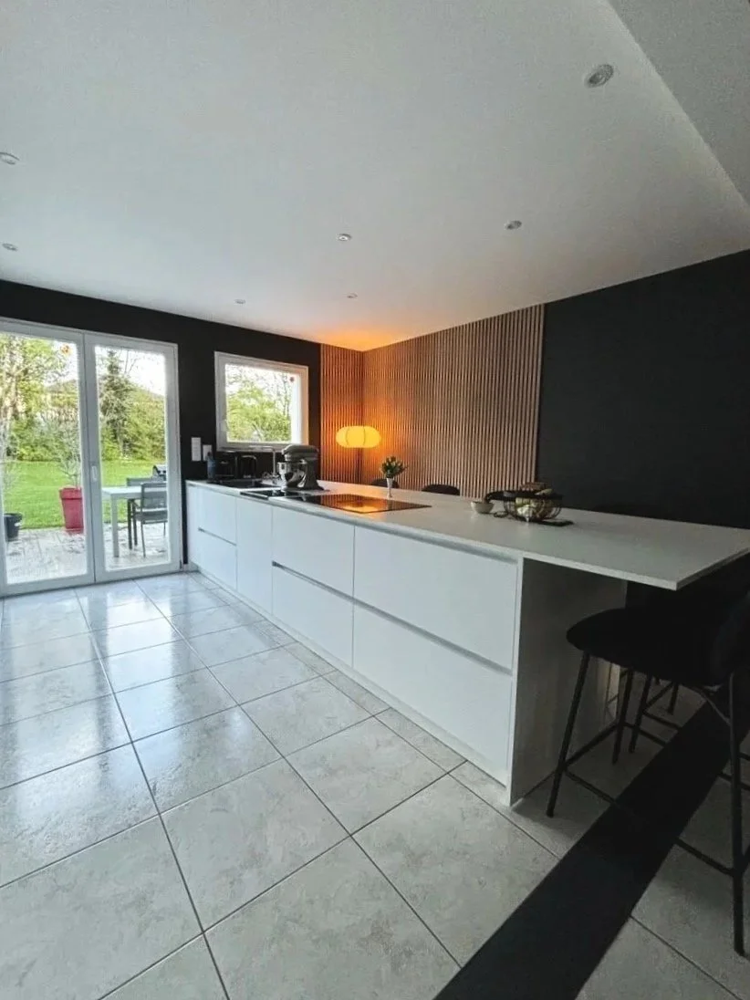 Cuisine moderne rénovée dans la maison à charnay avec plans de travail blancs, fenêtres donnant sur un jardin, lampadaire orange, murs noirs avec une paroi en bois, carrelage gris, quelques objets de cuisine et huit chaises noires.