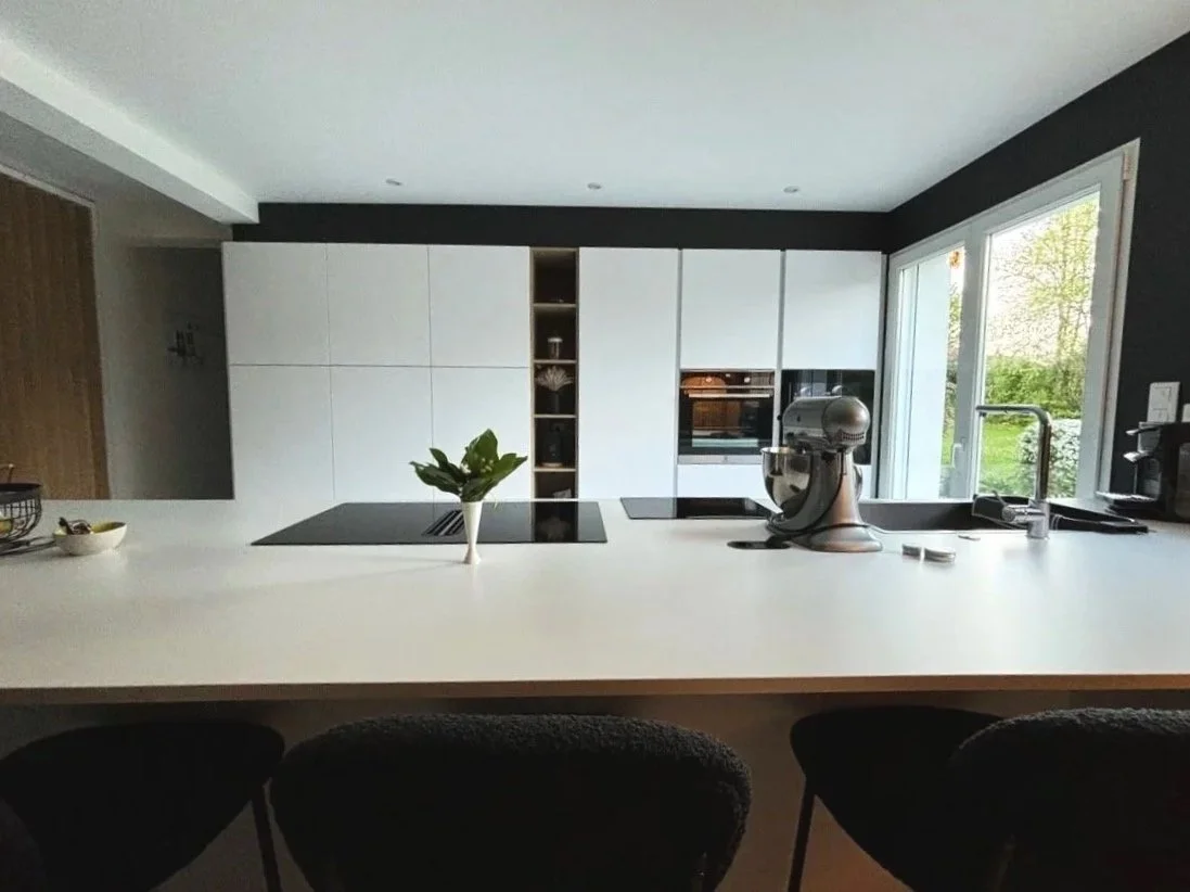 Cuisine moderne rénovée dans la maison à charnay avec des armoires blanches, un plan de travail blanc, une fenêtre donnant sur un jardin, un robot de cuisine sur le plan de travail, et des sièges noirs en premier plan.
