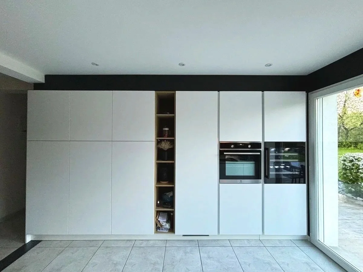 Cuisine moderne sur mesures rénovée dans la maison à charnay avec colonnes blanches, accents noirs et une grande fenêtre laissant entrer la lumière naturelle, donnant sur un espace extérieur verdoyant. 