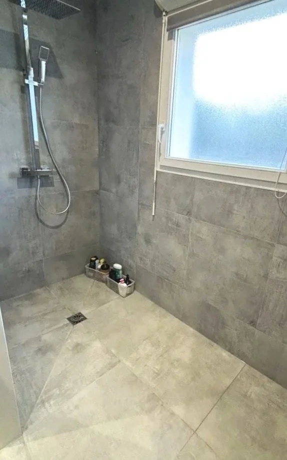 Salle de douche moderne avec grande fenêtre, paroi en carrelage gris, douches à main et étagère contenant divers produits de toilette, sol en carreaux gris, et un drain de sol. Agencement sur mesure. Maison charnay.