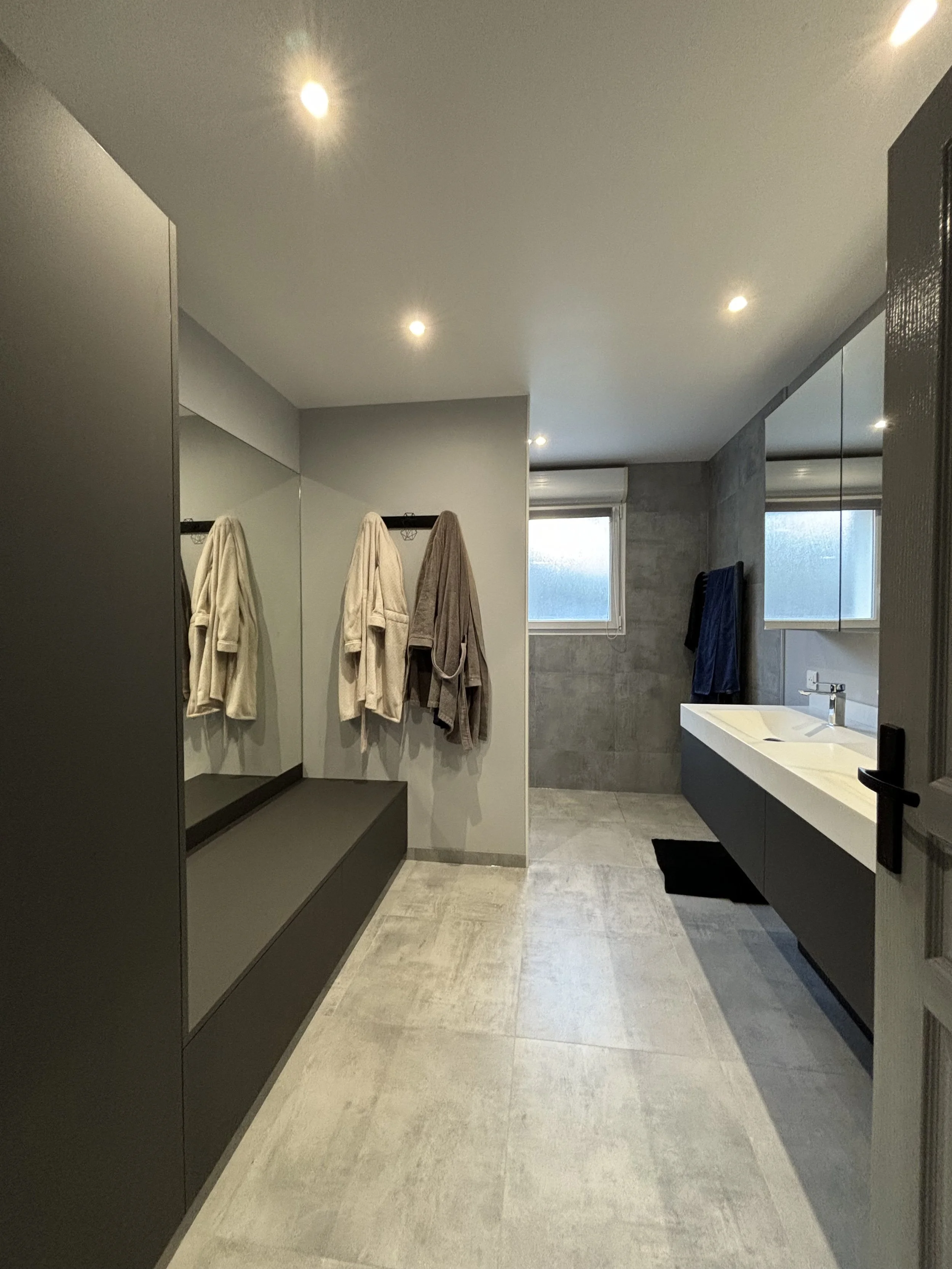 Salle de bain moderne avec baignoire, lavabo, miroir, fenêtre, et peignoirs suspendus.  Agencement sur mesure. Maison charnay.
