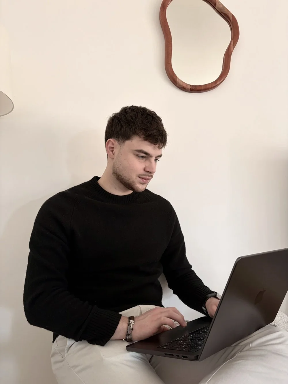 Jeune homme assis, utilisant un ordinateur portable en travaillant ses projets d'architecture d'intérieur, portant un pull noir et un pantalon beige, dans une pièce avec un mur blanc et un miroir design