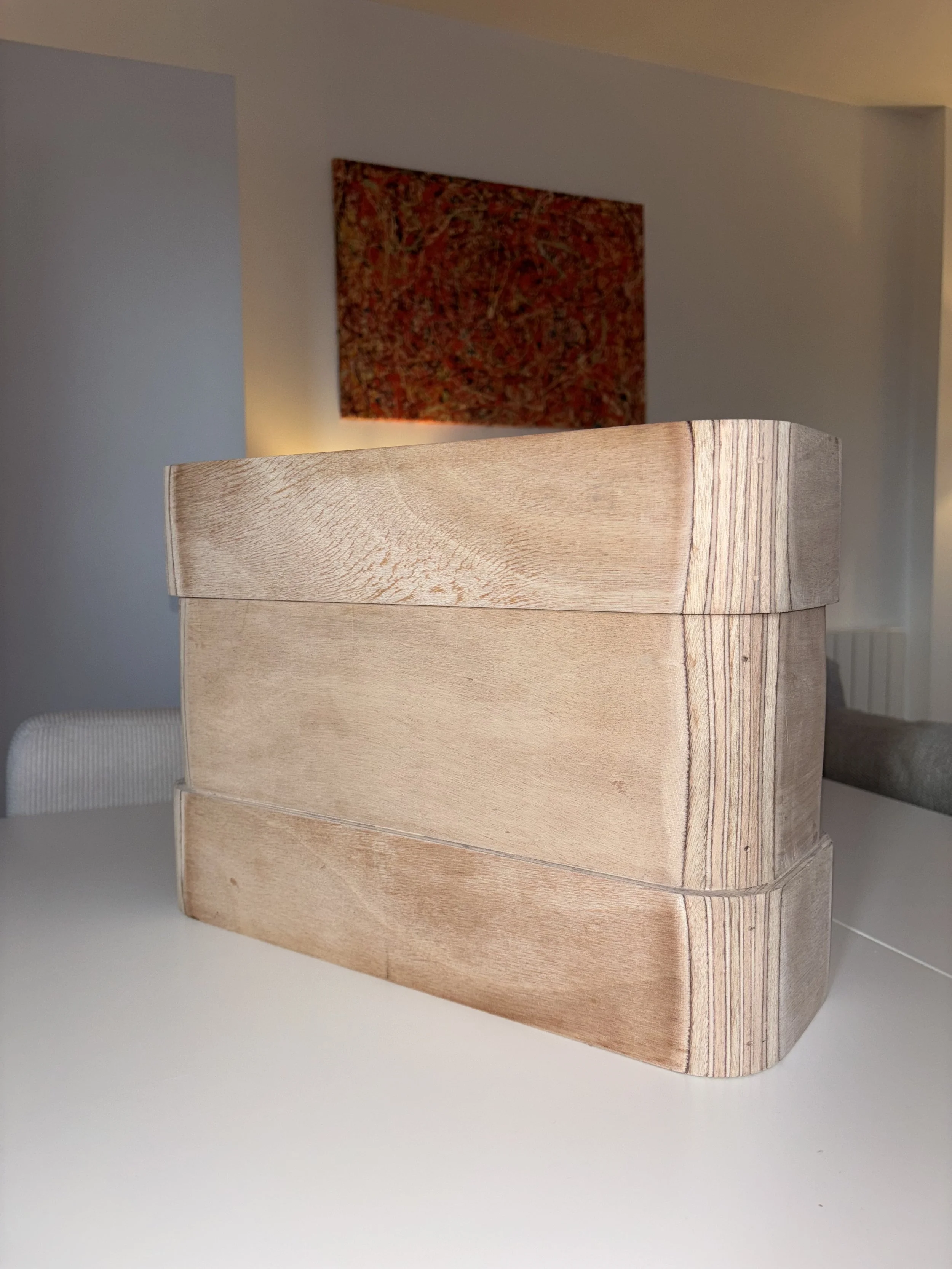 Un meuble en bois clair avec un motif incurvé en haut, placé sur une surface blanche, avec une peinture abstraite derrière.