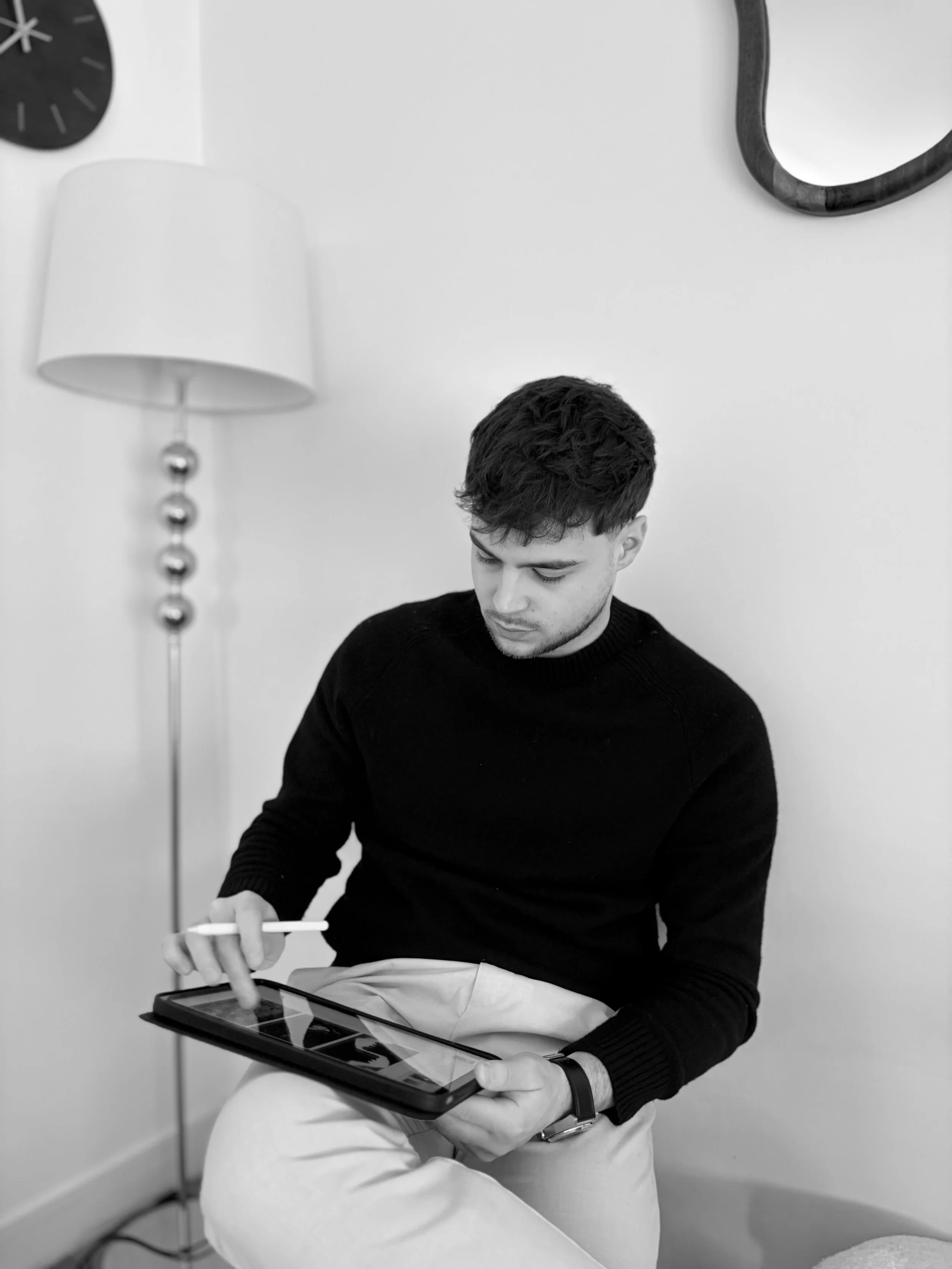 Lucas Frachebois, un jeune architecte d'intérieur, portant un pull noir et une montre noire, utilisant une tablette numérique avec un stylet qui fait ses projets d'architecture d'intérieur, avec un mur blanc derrière lui.