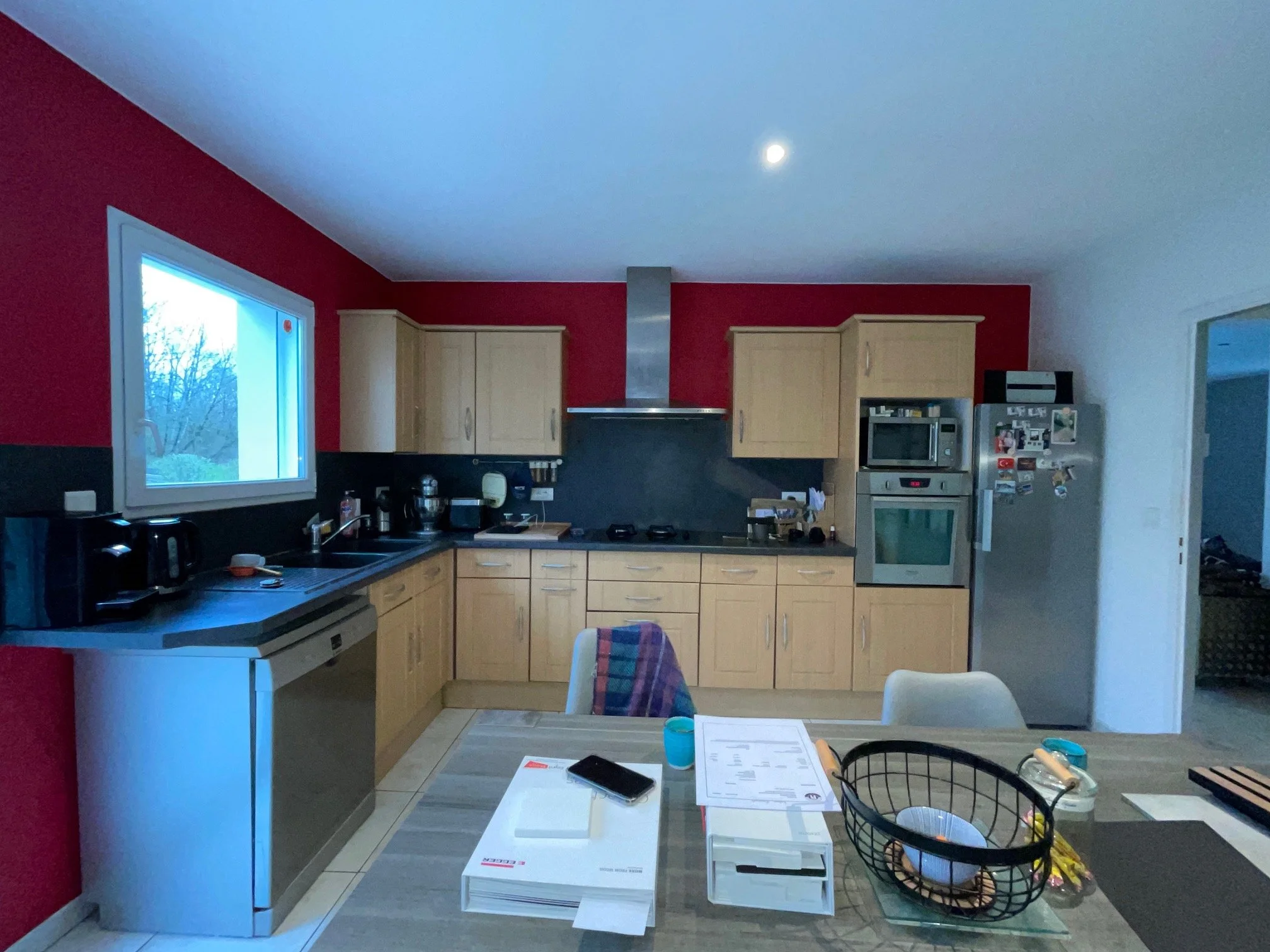 Cuisine existante de la maison rénovée à charnay avec murs rouges, armoires en bois, un plan de travail noir, deux fenêtres donnant sur un jardin et une porte-fenêtre.