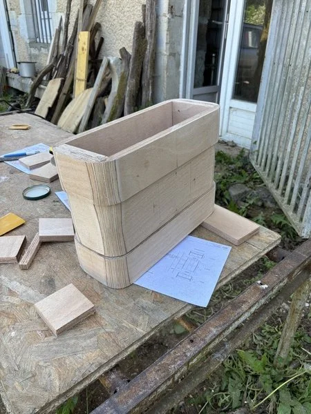 Une pièce en bois en cours de fabrication sur un établi en bois, avec des plans de construction et des morceaux de bois autour, en extérieur.