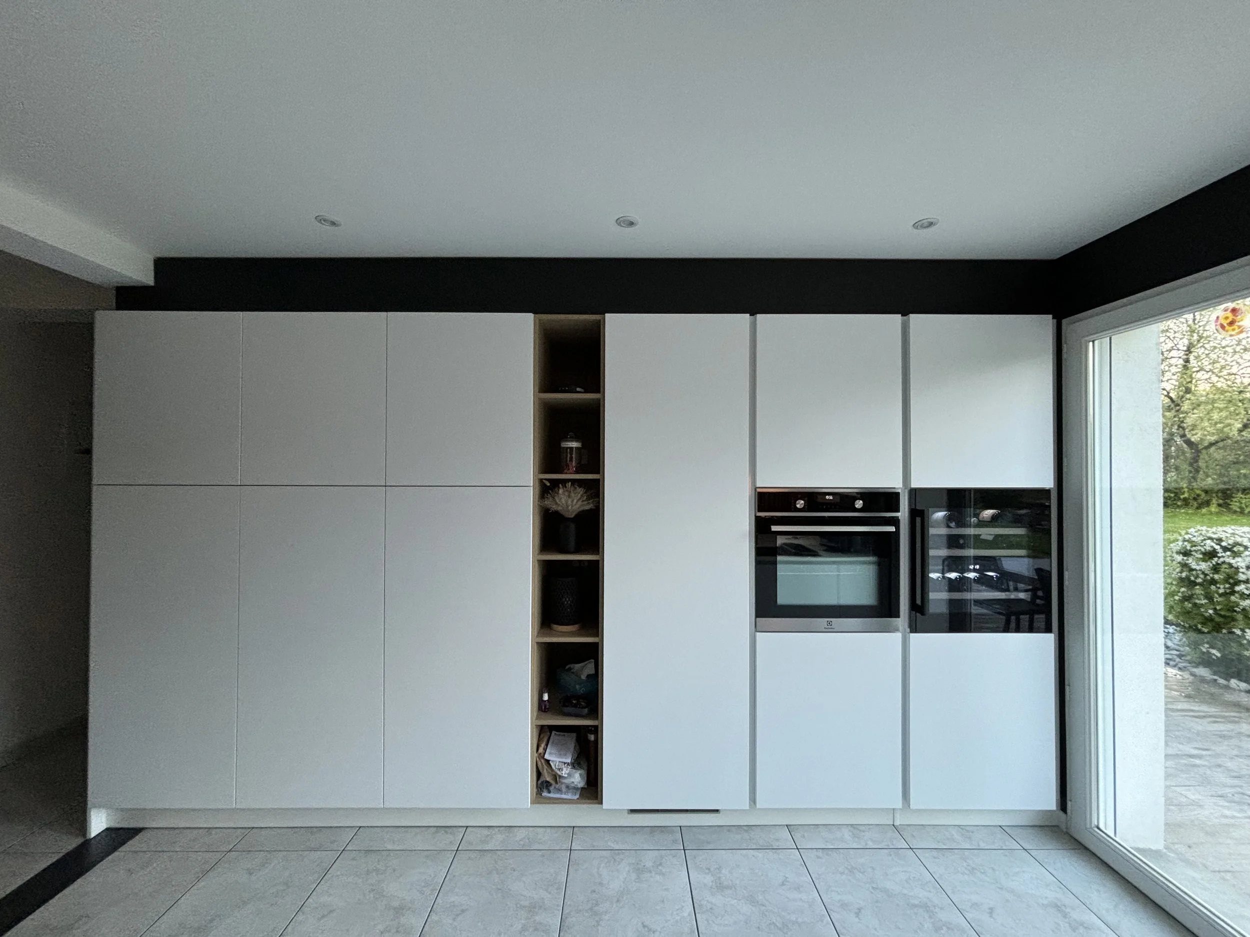 Cuisine moderne sur mesures rénovée dans la maison à charnay avec colonnes blanches, accents noirs et une grande fenêtre laissant entrer la lumière naturelle, donnant sur un espace extérieur verdoyant. 