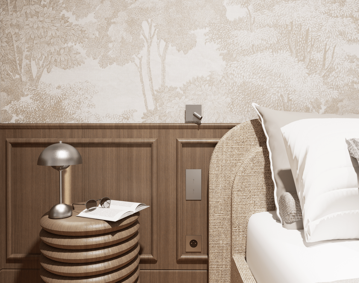 Coin de chambre avec une table en bois rond, une lampe de bureau, des lunettes, un livre ouvert, un lit avec des oreillers, un panneau en bois sur le mur avec une tapisserie ARTE de forêt beige en fond et une tête de lit en rotin tissé.