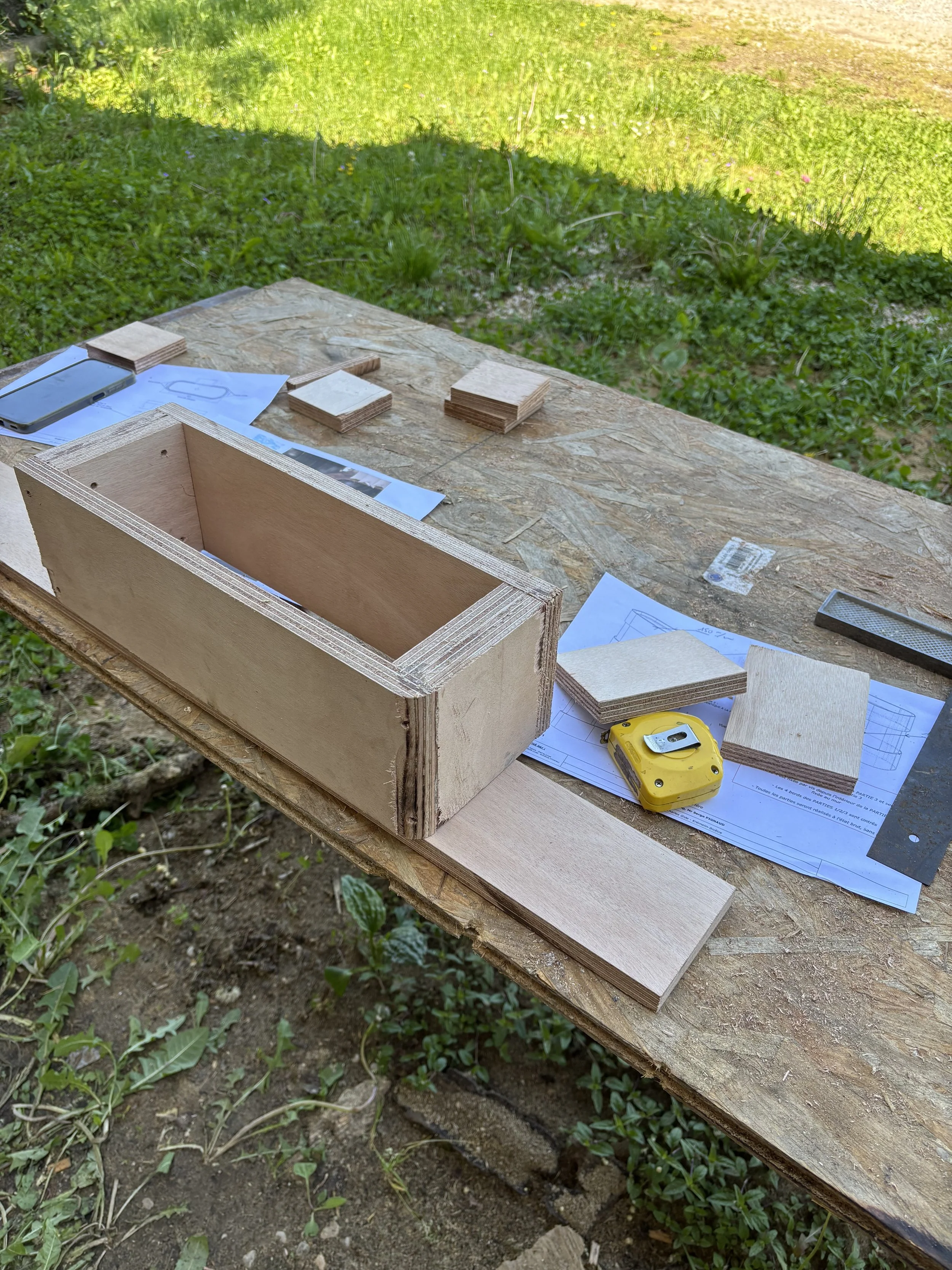 Table de travail en bois avec des morceaux de bois, des plans, un mètre ruban jaune, et un téléphone portable, extérieur sur une surface en bois avec de l'herbe verte à l'arrière.