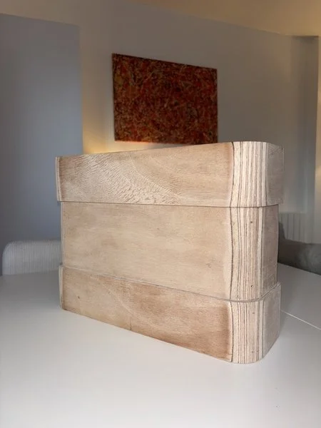 Un meuble en bois clair avec un motif incurvé en haut, placé sur une surface blanche, avec une peinture abstraite derrière.