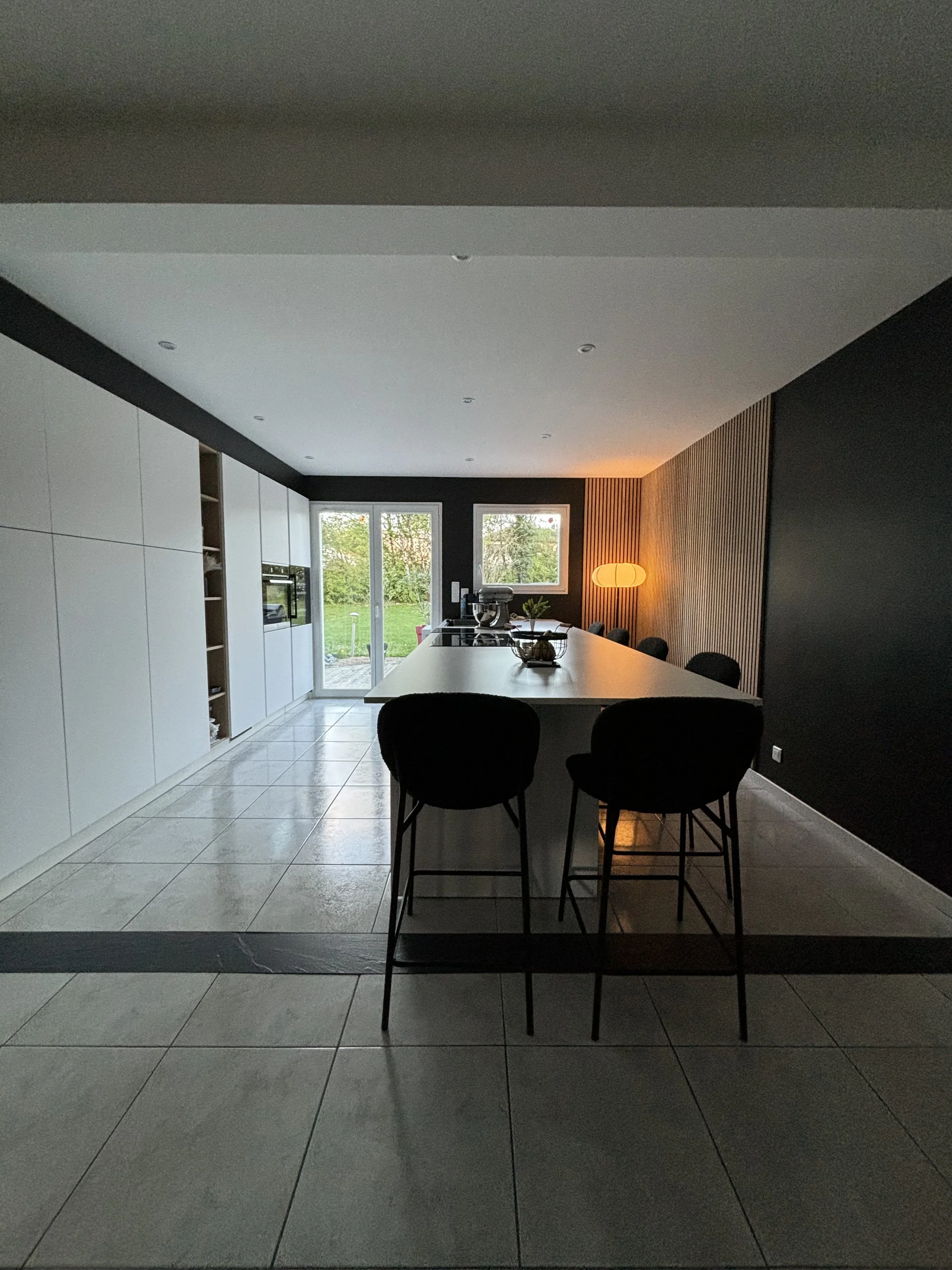 Cuisine rénovée dans la maison à charnay moderne avec un îlot central, chaises, grande fenêtre laissant entrer de la lumière naturelle, mur en bois, lampe sur le mur à droite.