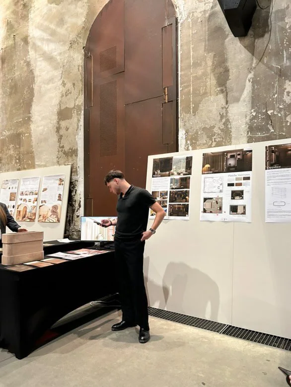 Jeune homme regardant son téléphone dans une salle d'exposition avec des affiches et des plans sur le mur, murs en pierre et porte métallique en arrière-plan.