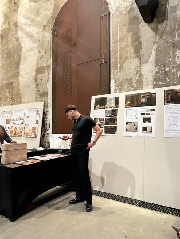 Jeune homme regardant son téléphone dans une salle d'exposition avec des affiches et des plans sur le mur, murs en pierre et porte métallique en arrière-plan.