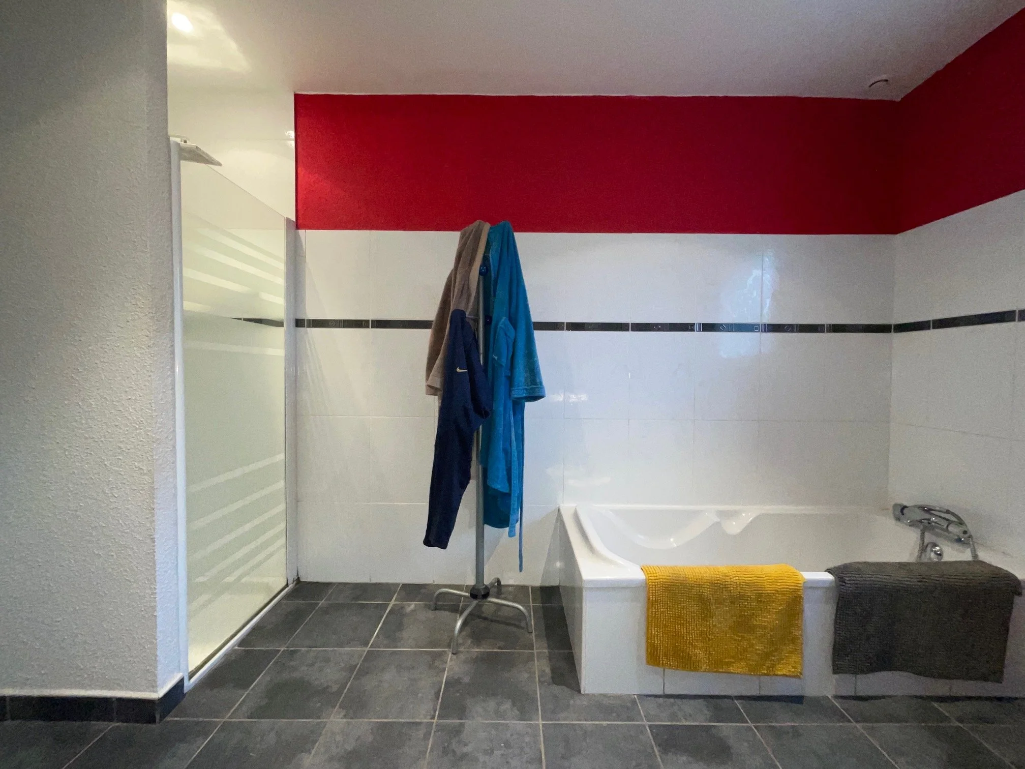 Salle de bain existante de la maison rénovée à charnay avec baignoire, miroir, porte de douche en verre, étendoir avec vêtements suspendus, serviette jaune sur la baignoire, mur rouge et blanc, carrelage noir et blanc, sol noir.