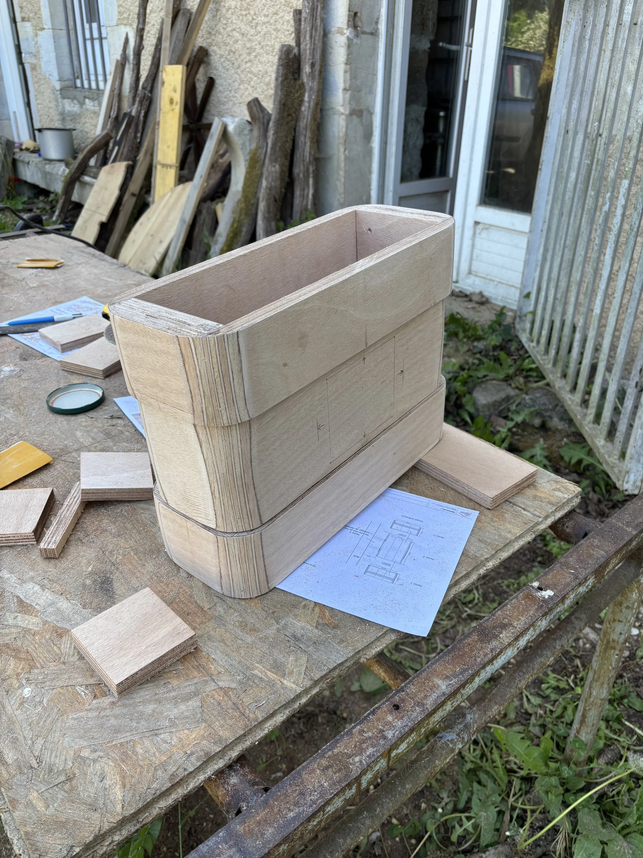 Une pièce en bois en cours de fabrication sur un établi en bois, avec des plans de construction et des morceaux de bois autour, en extérieur.