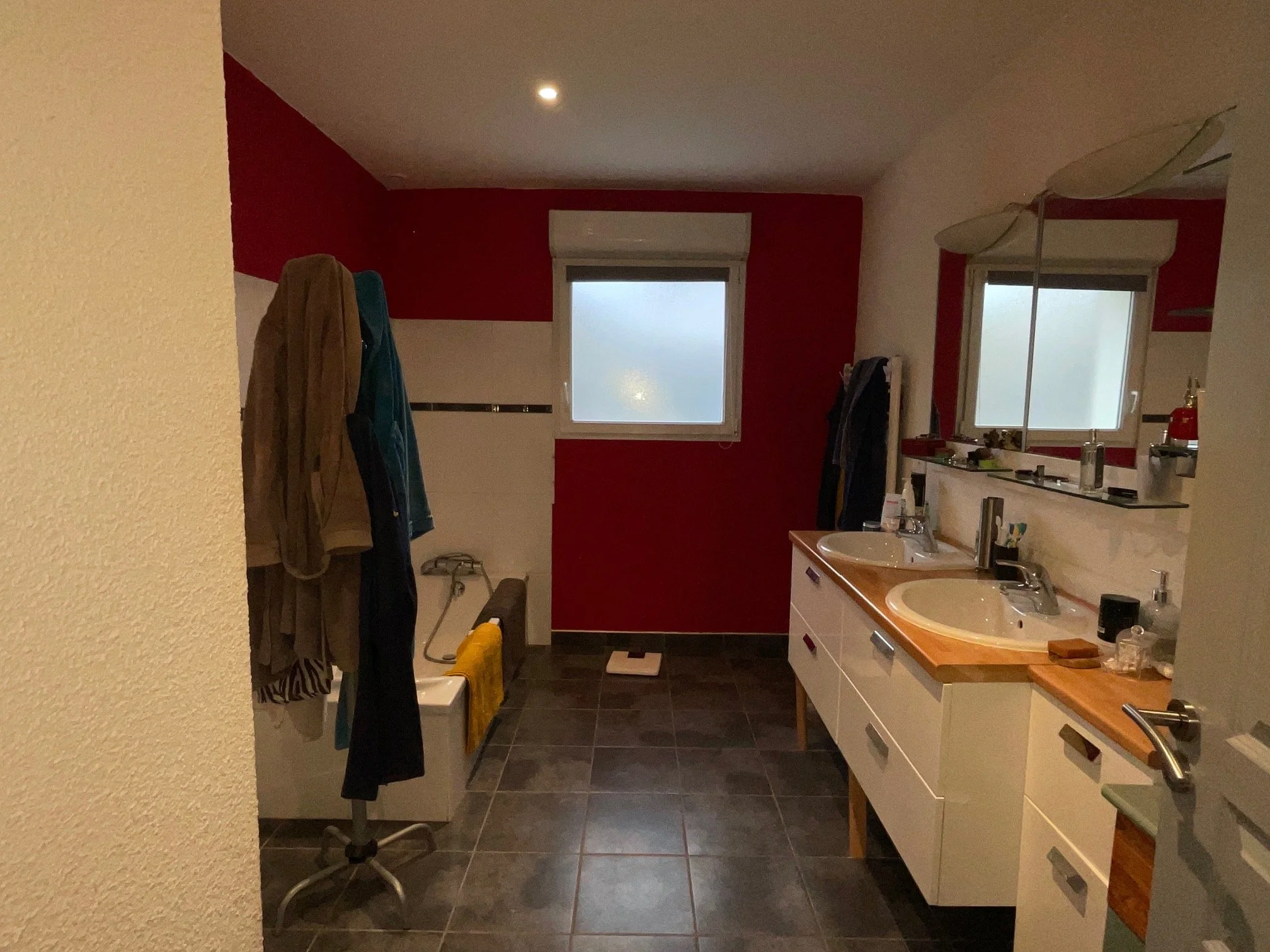 Salle de bain existante de la maison rénovée à charnay avec deux lavabos, des miroirs, une fenêtre, un sèche-serviettes et des vêtements suspendus. Le mur derrière les lavabos est rouge, le sol est en carreaux noirs.