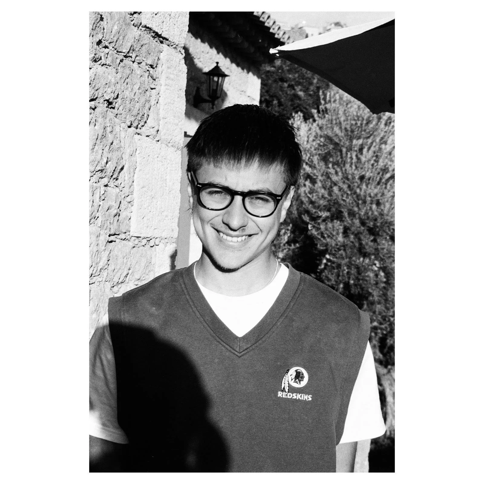 Black and white photo of a young man with glasses smiling, wearing a Redskins sports jersey, standing outdoors next to a stone wall with trees and a building in the background.