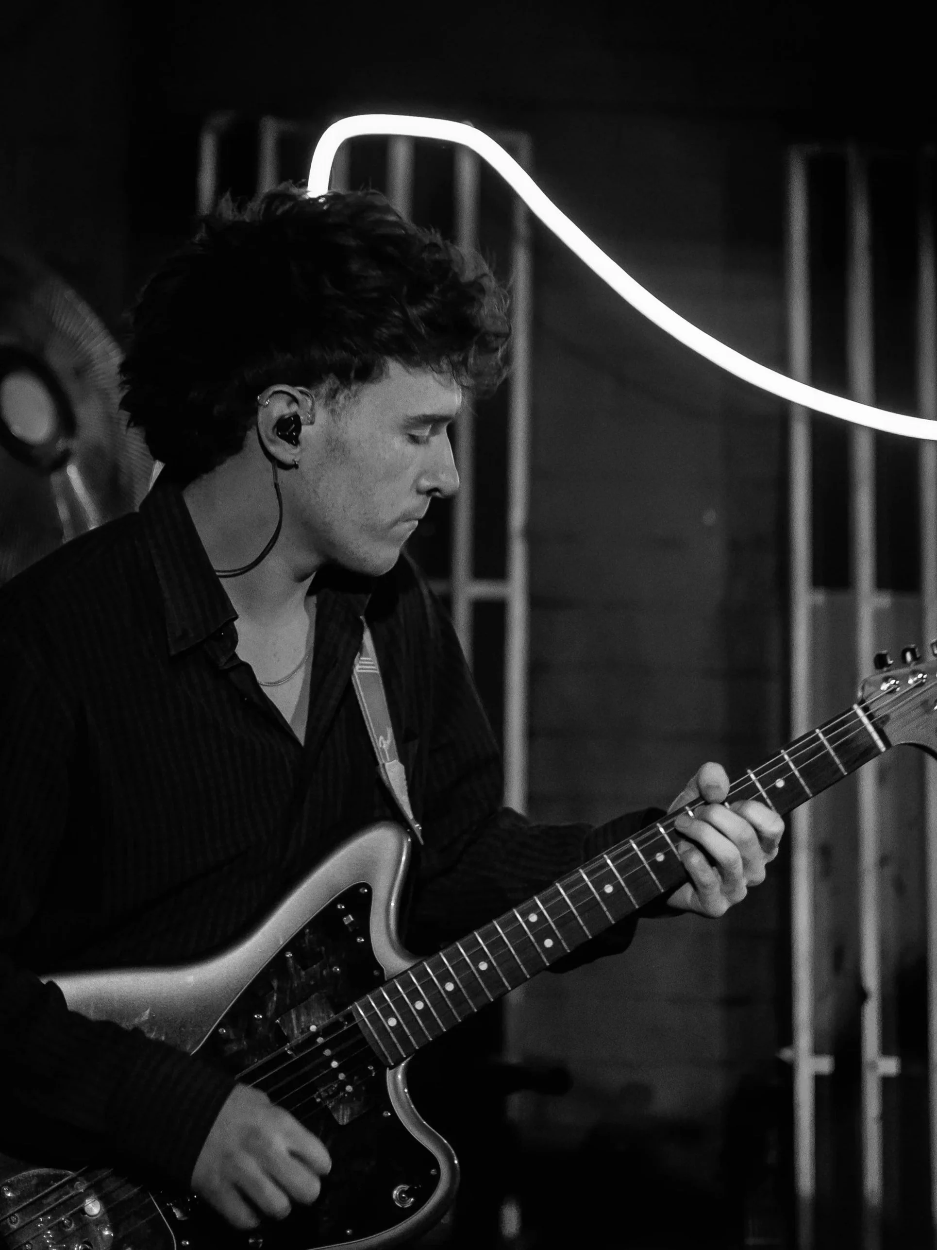 A young man with curly hair playing an electric guitar, wearing in-ear monitors, in a dimly lit indoor setting.