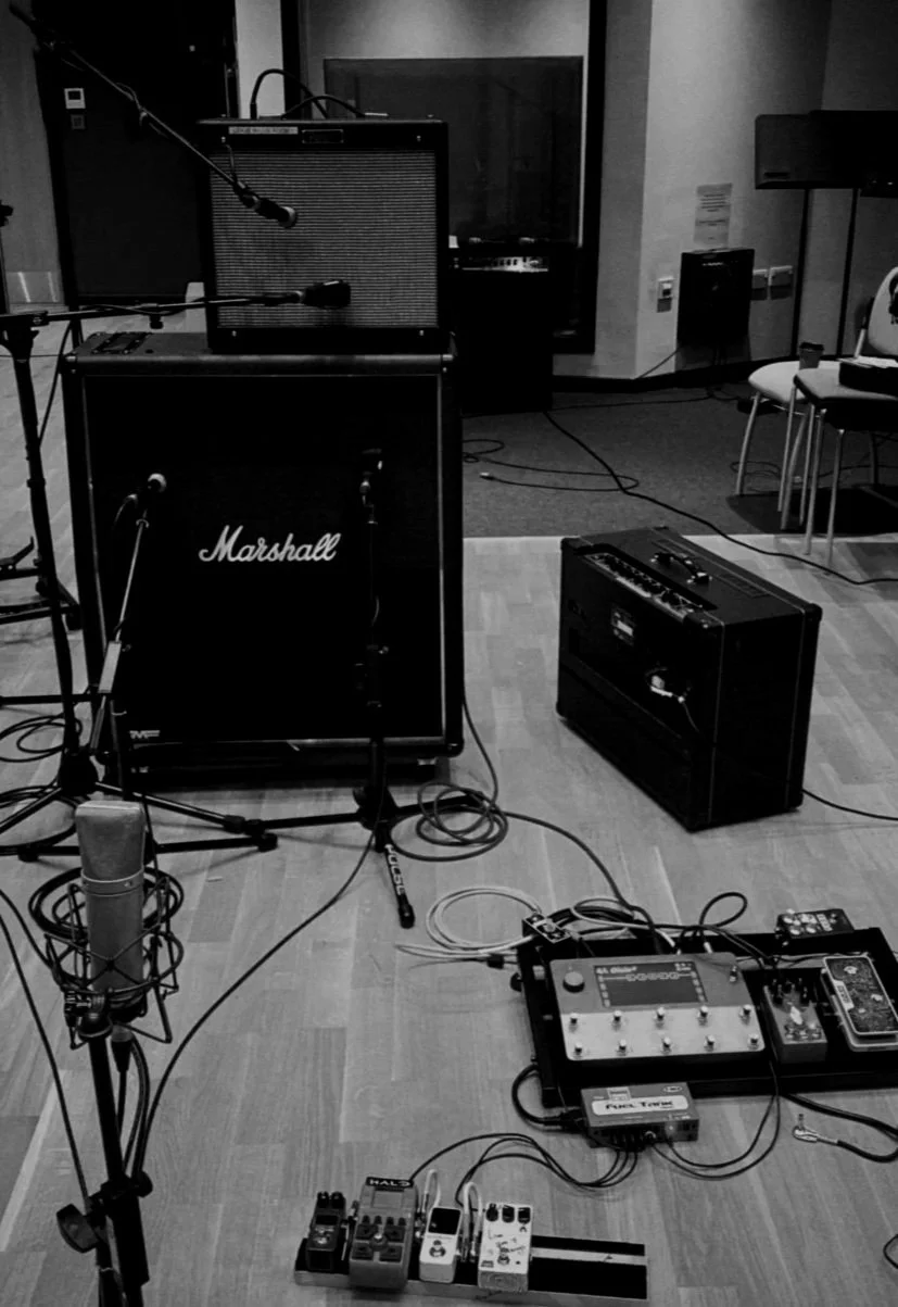 Musical recording studio setup with microphone, amplifiers (including a Marshall amp), and various effects pedals and equipment on the floor.