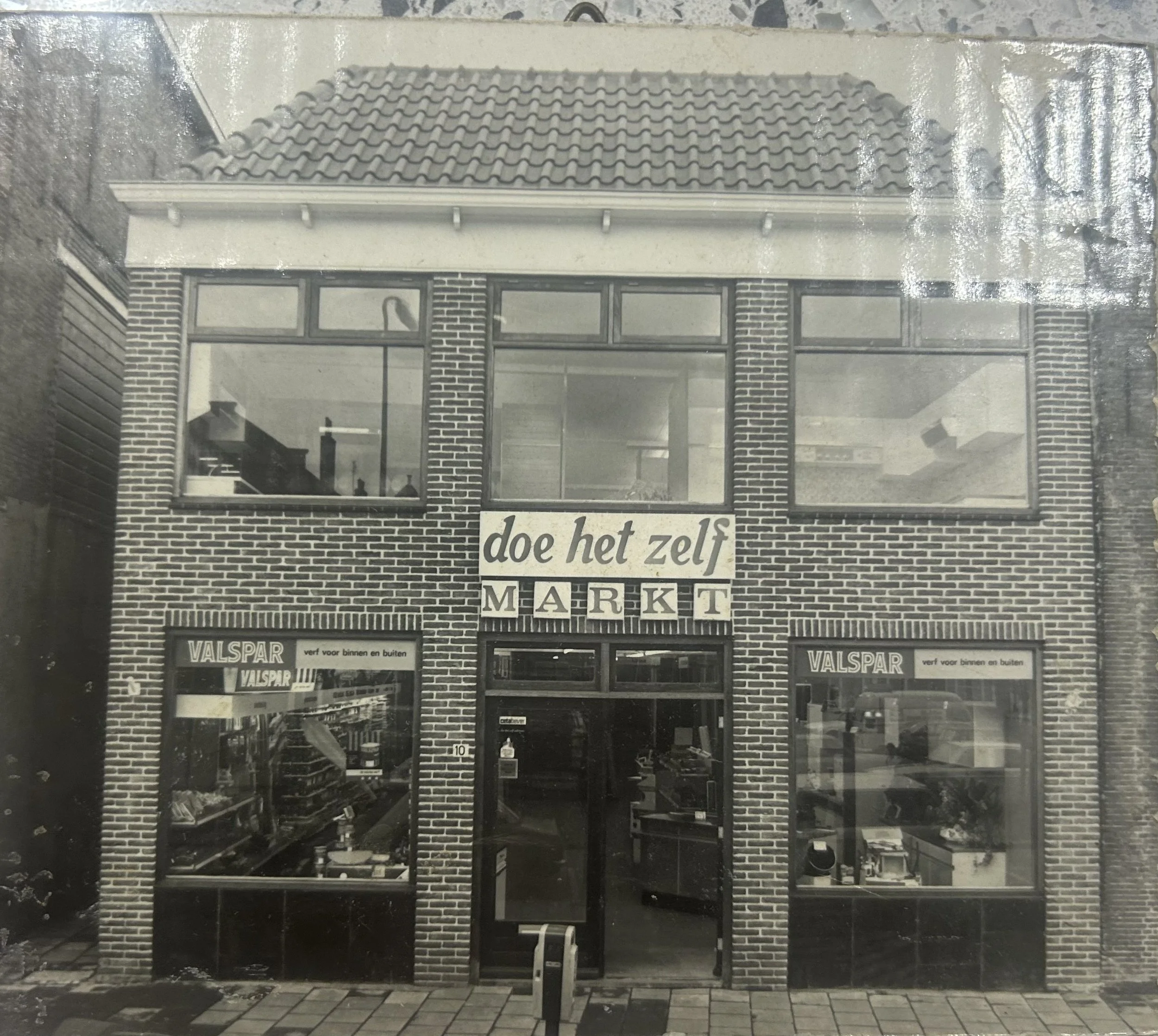Black and white photo of the facade of a brick store with large windows and a sign that reads 'do het zelf' and 'MARKT'. There are signs in the windows that read 'VALSPAR' and advertisements for paint.