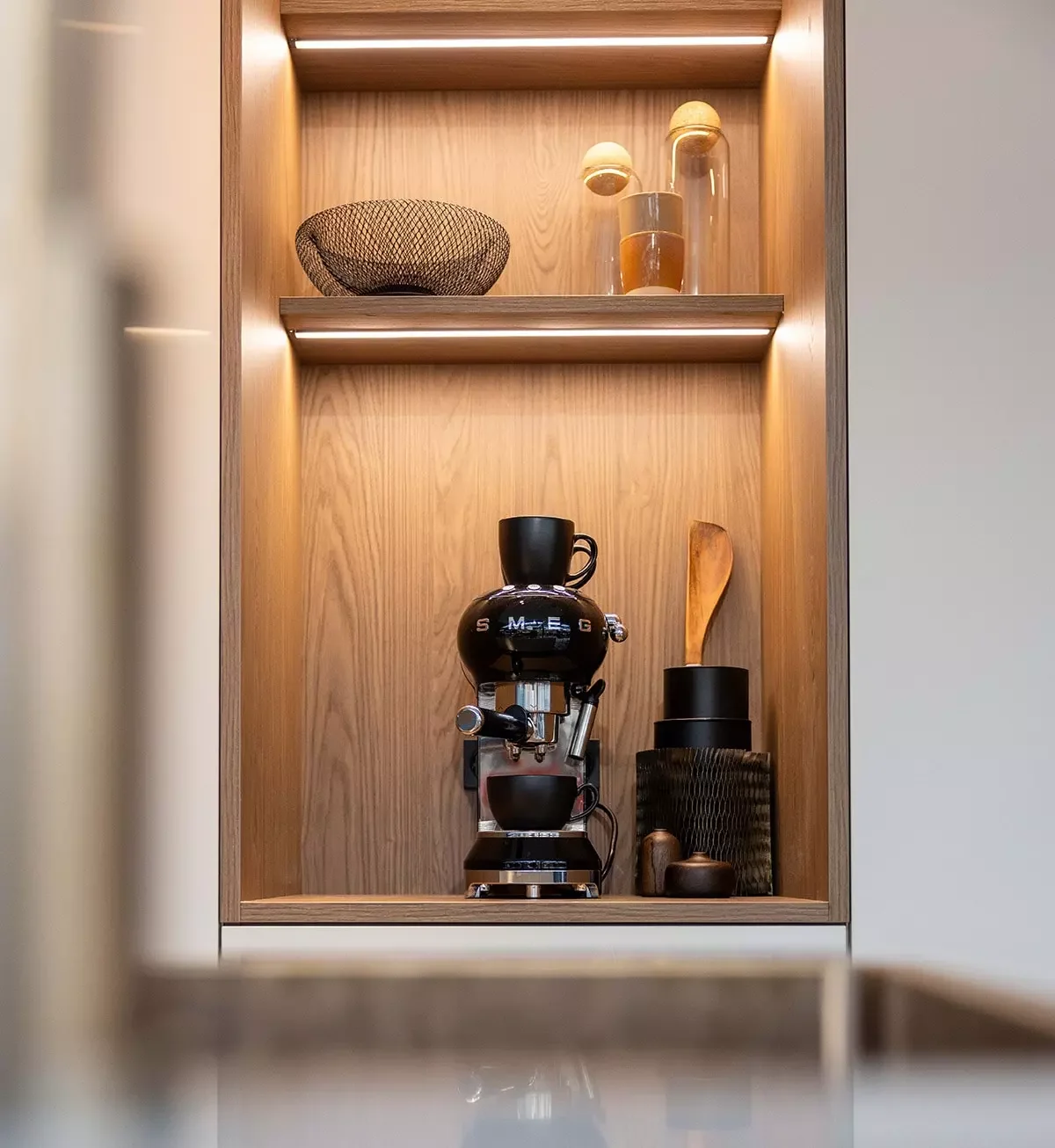 A wooden kitchen cabinet with a top shelf and a middle shelf. The top shelf holds a black wire bowl and two glass vases with decorative objects inside. The middle shelf contains a black coffee machine with a glass cup on top, a black container with a wooden spatula, and small decorative objects.