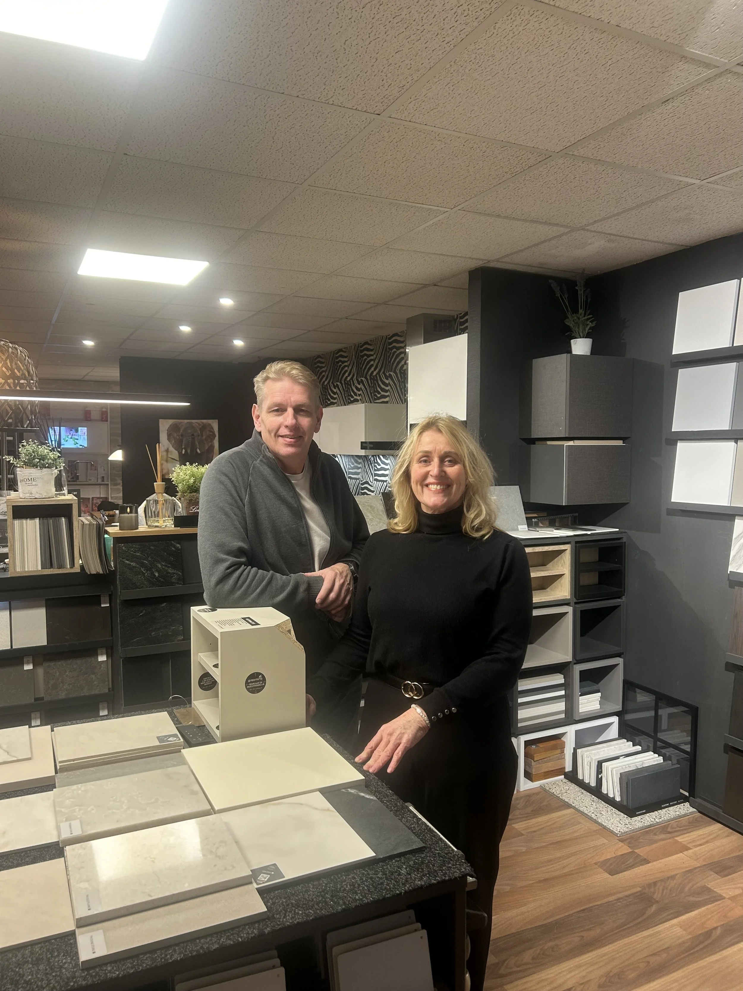 Two smiling women standing behind a display table in a showroom with various tile samples, shelves, and home decor items in the background.