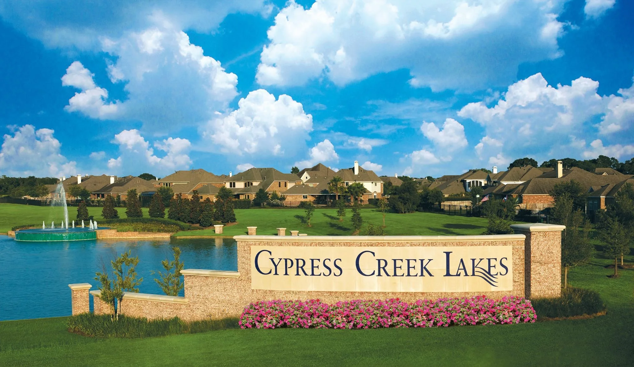 Entrance sign for Cypress Creek Lakes with houses, a water fountain, green lawn, and a blue sky with clouds in the background.